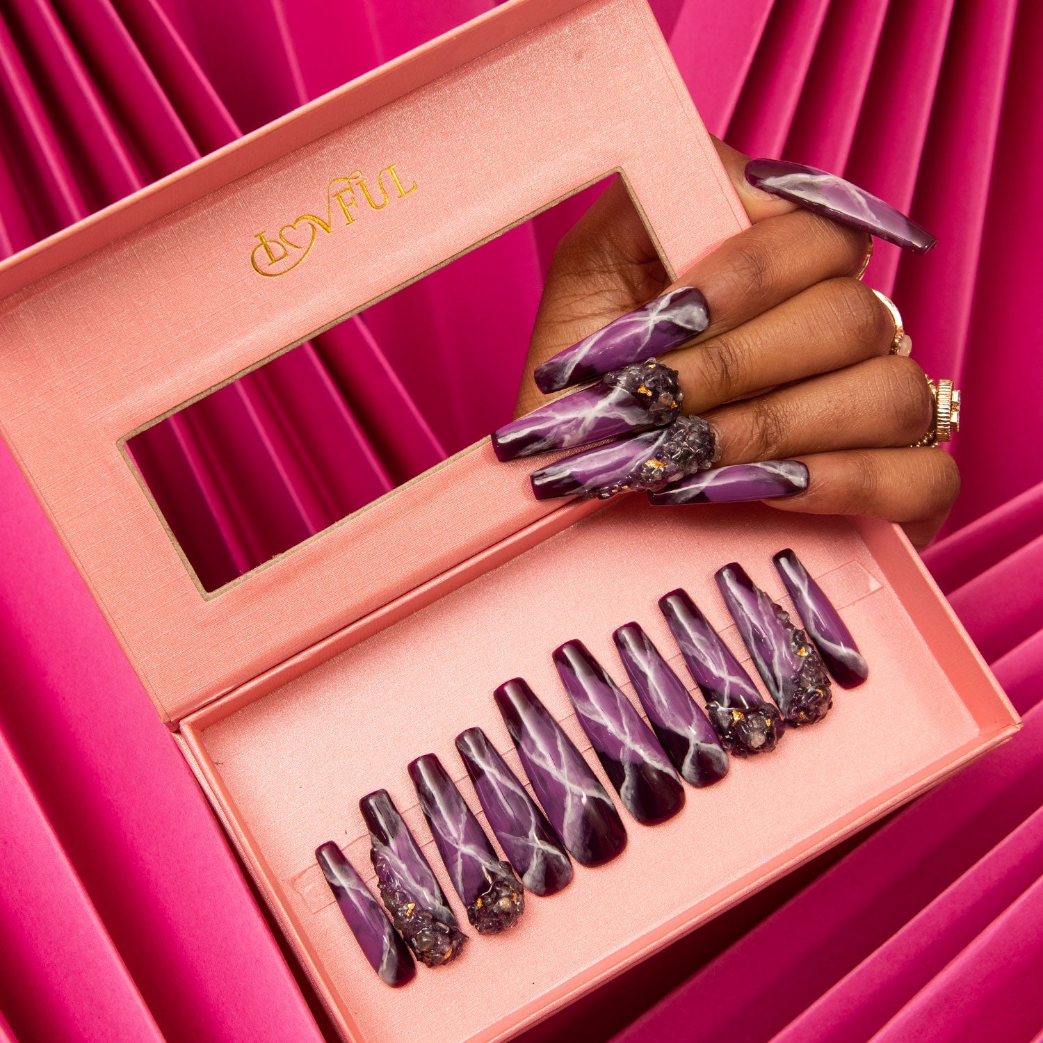 Dream Amethyst press-on coffin nails featuring purple and white swirling patterns and detailed embellishments, displayed in a pink Lovful box with a transparent window on a vibrant pink pleated fabric background.