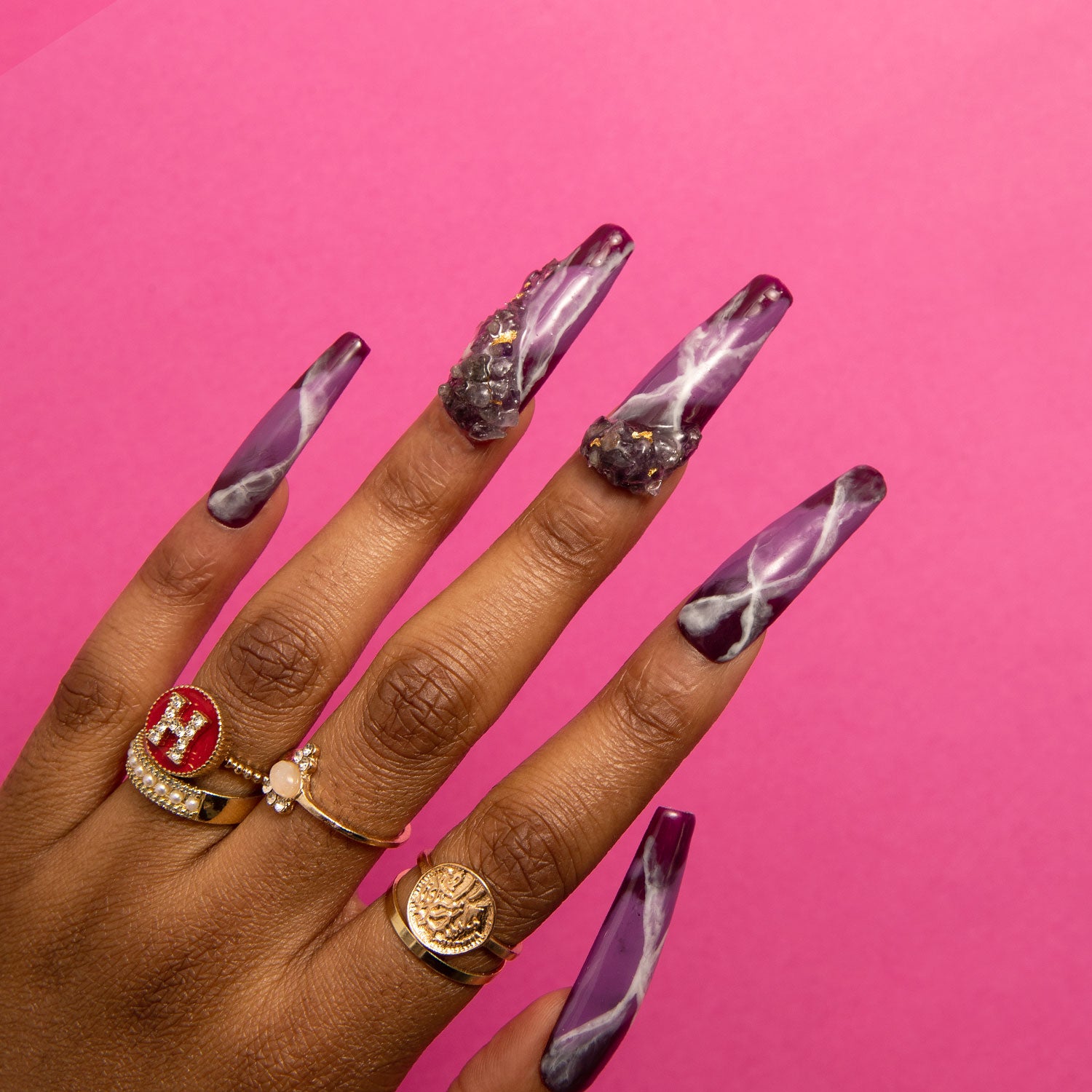 Hand showcasing Dream Amethyst Coffin press-on nails in shades of purple and white with marbled patterns against a pink background. The nails include intricate designs, perfect for adding a touch of mystique to any occasion.