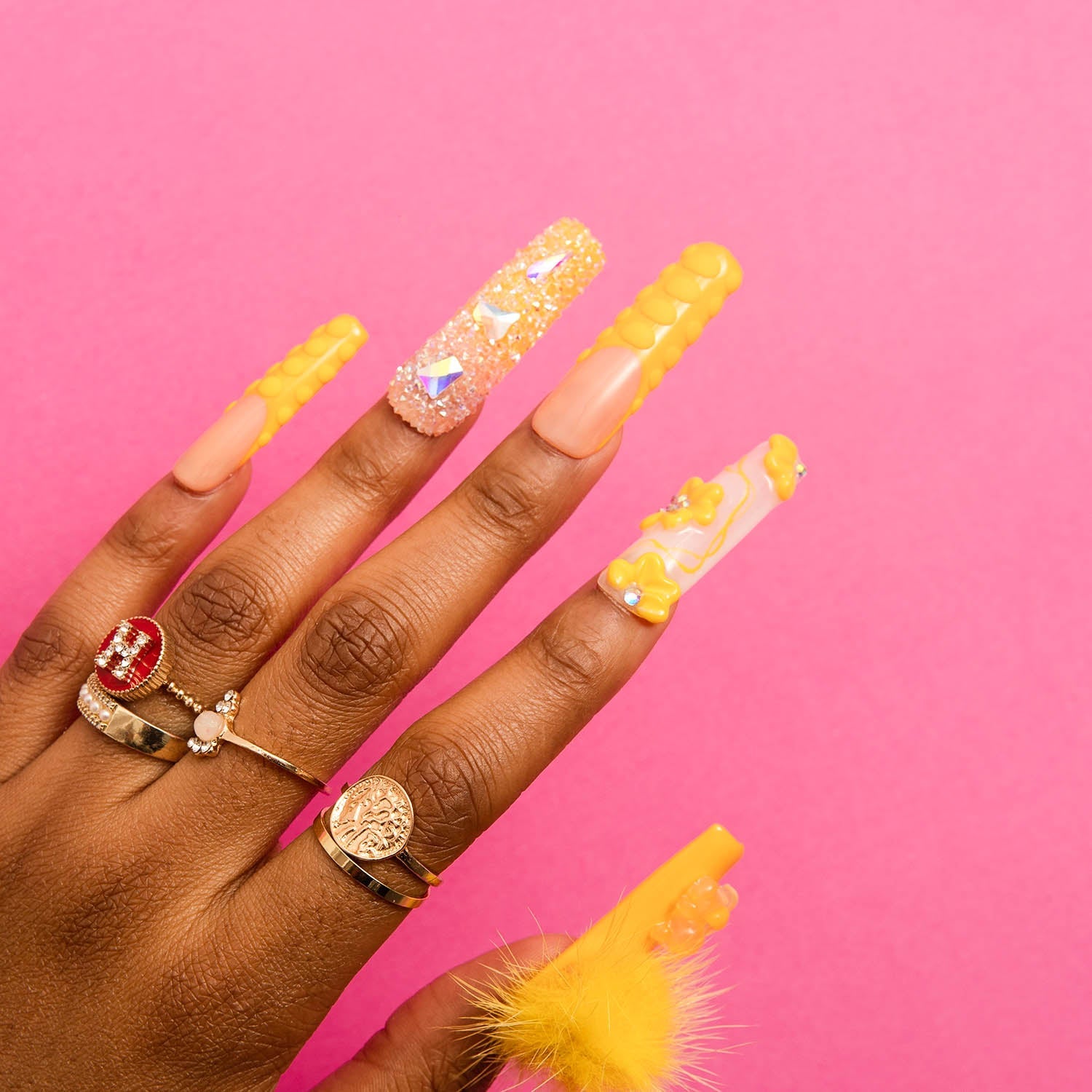 Hand with long, square-shaped press-on acrylic nails in sunny yellow, glittery, and 3D designs by Lovful. A detachable fluffy ball adorns one nail. Hand wears statement rings, set against a bright pink background.