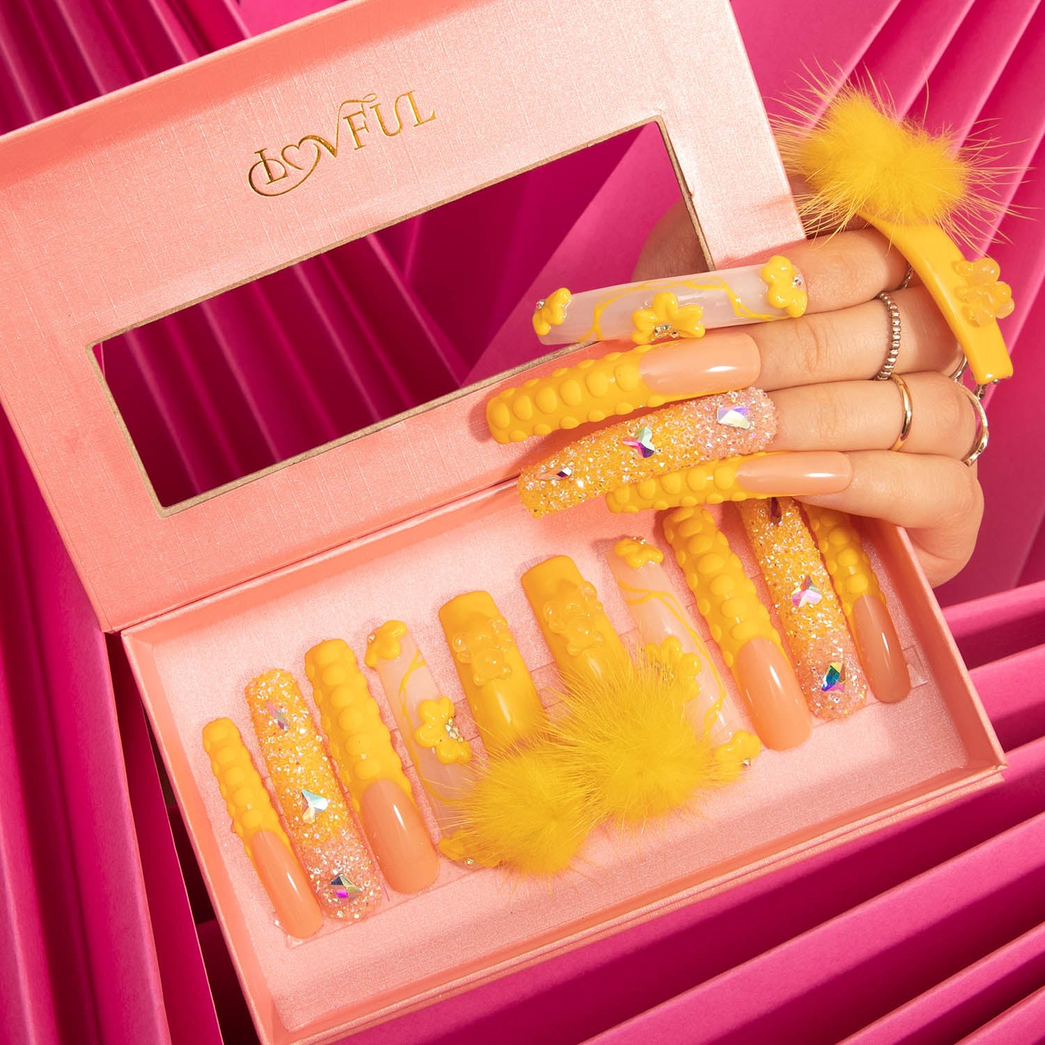 Lovful 'Yummy' press-on nails from the H131 collection in a pink box, featuring sunny yellow hue with detachable fluffy yellow balls, glitter, and textured patterns. A hand displays the applied nails.