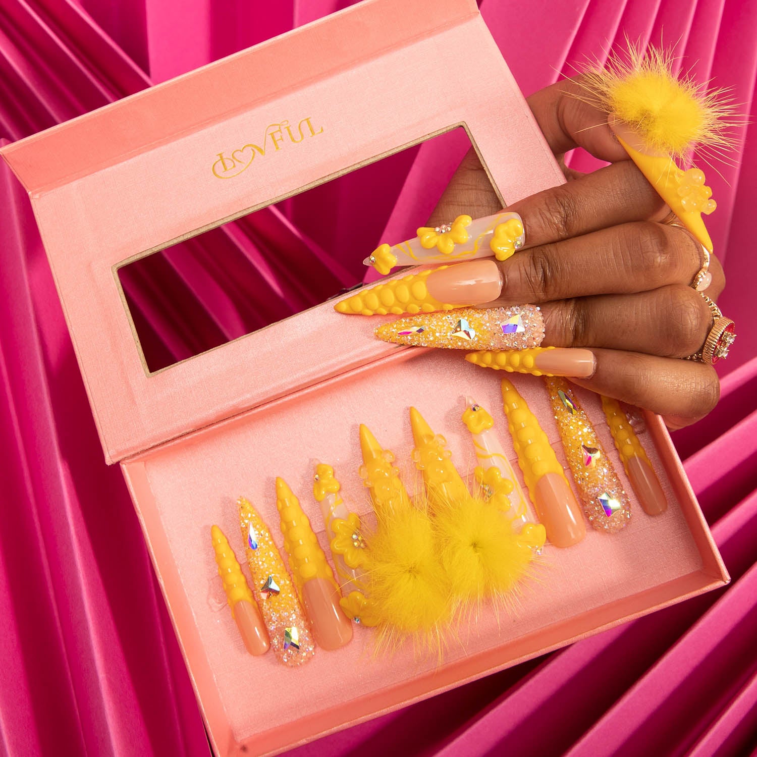 Hand displaying sunny yellow stiletto press-on nails with detachable fluffy balls and decorative glitter elements in a pink Lovful branded box.