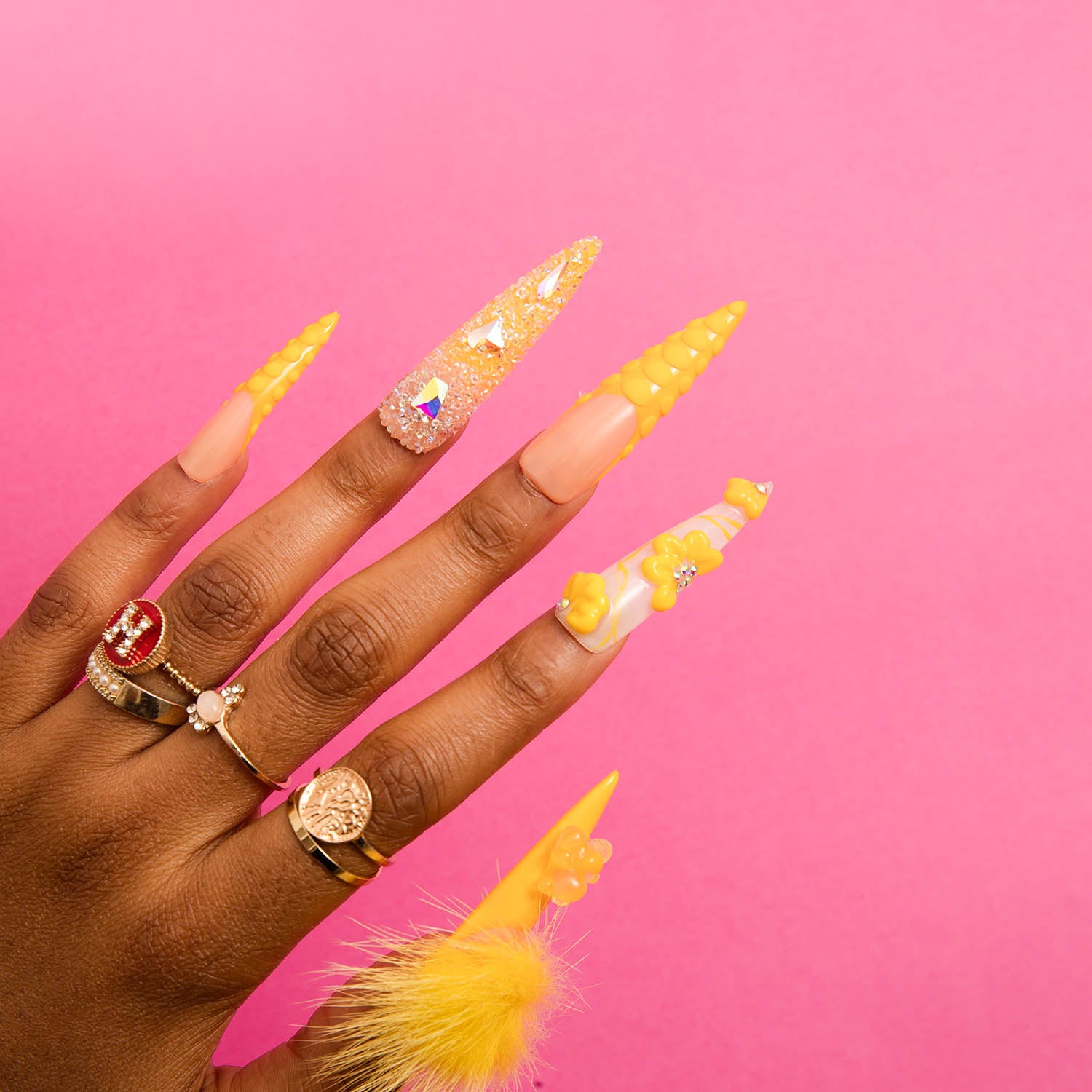 Hand with stiletto-shaped, sunny yellow acrylic nails featuring glitter, beads, flowers, and a detachable fluffy ball, highlighting the Yummy nail design from Lovful on a pink background.