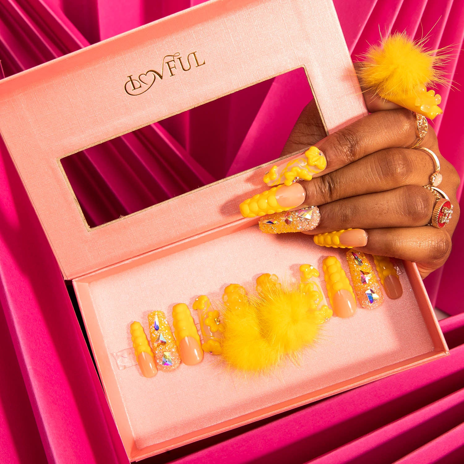 Hand showcasing Lovful's Yummy Coffin press-on nails with detachable fluffy balls and glitter, in a pink box against a pink background. Perfect for adding a playful twist to your summer look.