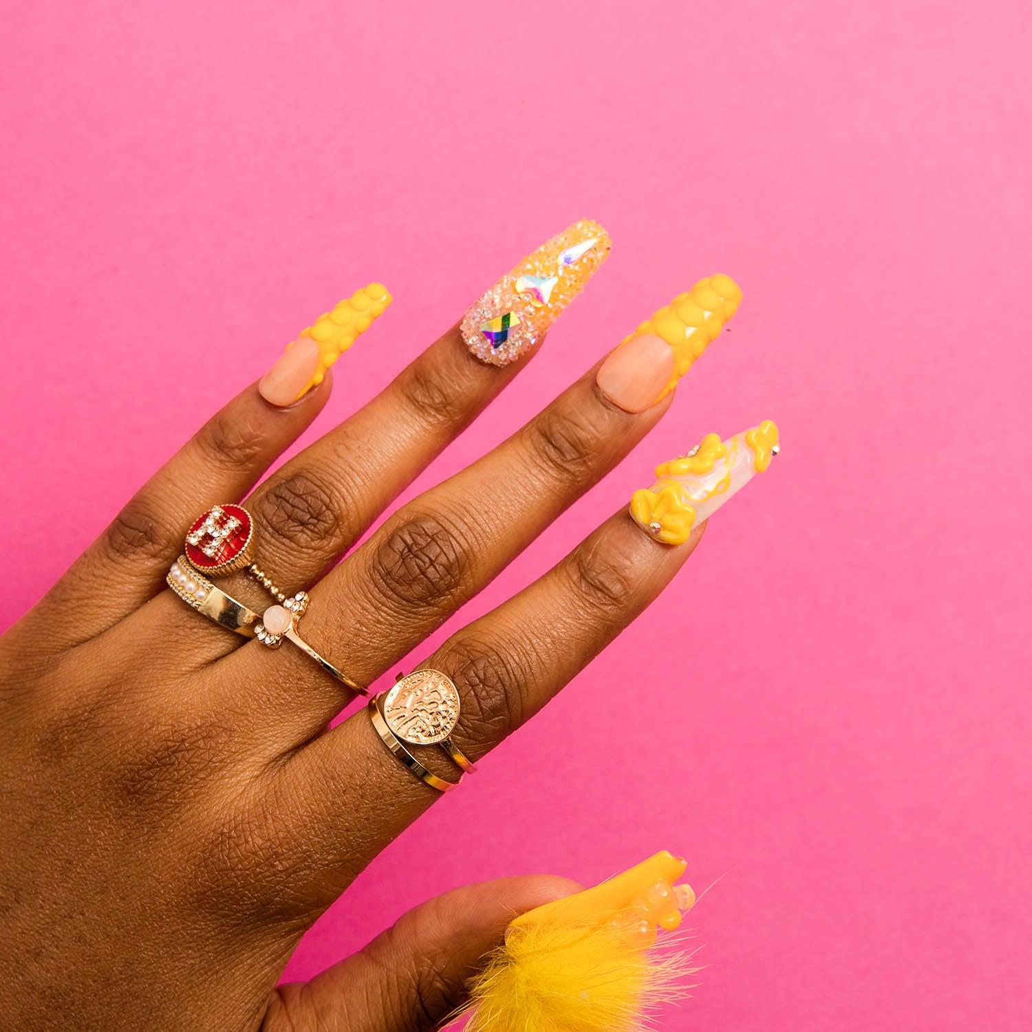 Hand showcasing yellow coffin-shaped press-on acrylic nails with golden embellishments and fluffy yellow balls on a pink background, accessorized with multiple rings.