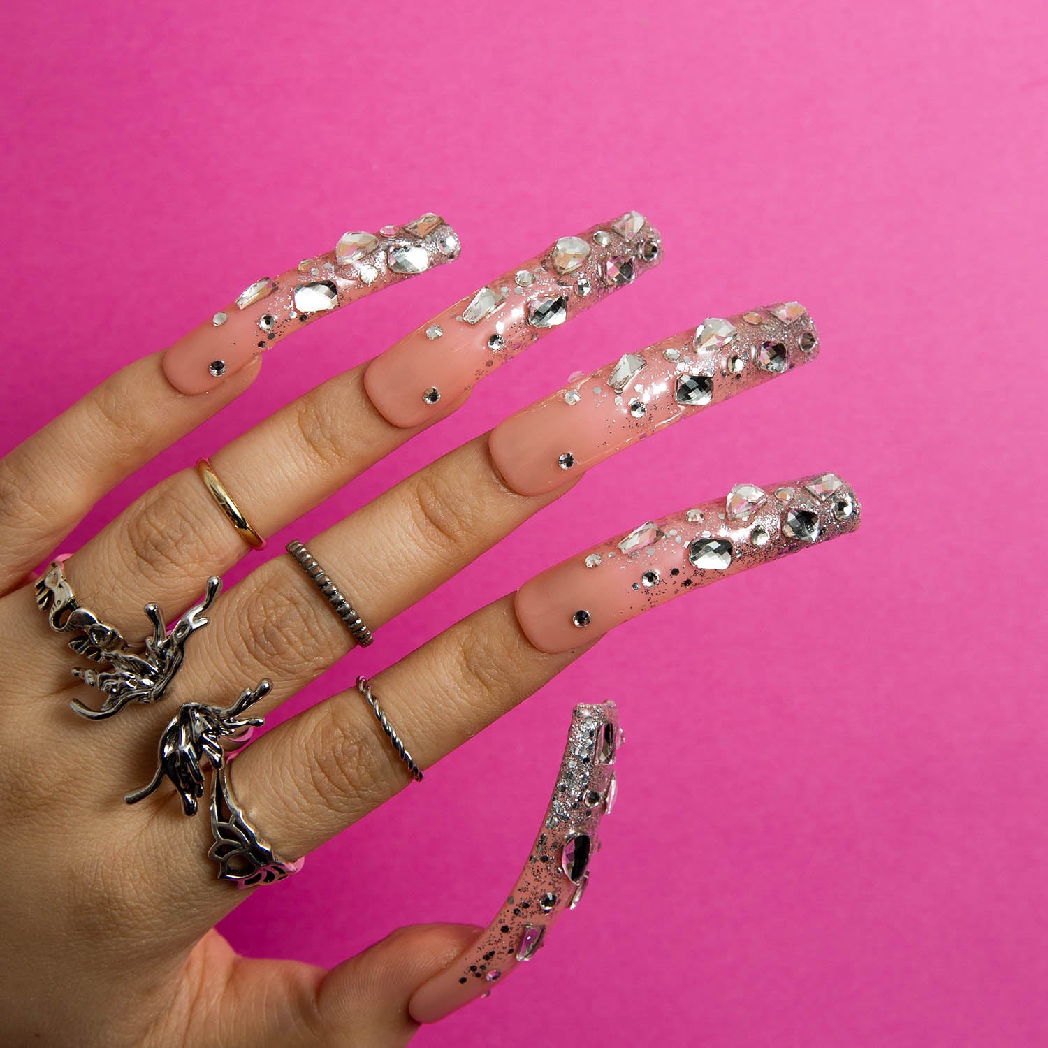 Hand with long press-on acrylic nails featuring silver glitter and sparkling rhinestones, against a pink background. The nails have a curved shape, and the hand is wearing multiple rings. 'Silver Wind' design by Lovful.