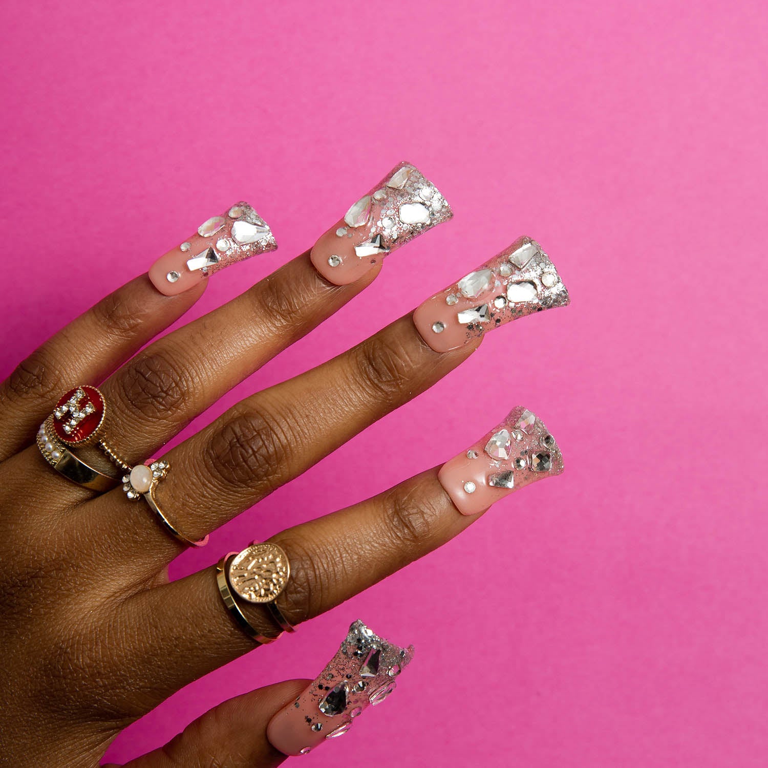 Hand with long square press-on nails called 'Silver Wind', featuring silver glitter and sparkling rhinestones, against a pink background. Several gold and silver rings are worn on the fingers.