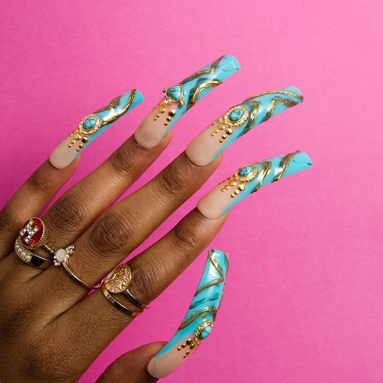 Hand with long, curved turquoise press-on acrylic nails adorned with golden decor and sparkling gems, against a pink background. The nails feature intricate designs, and the hand wears various rings including a red gemstone ring, a pearl ring, and a gold signet ring.