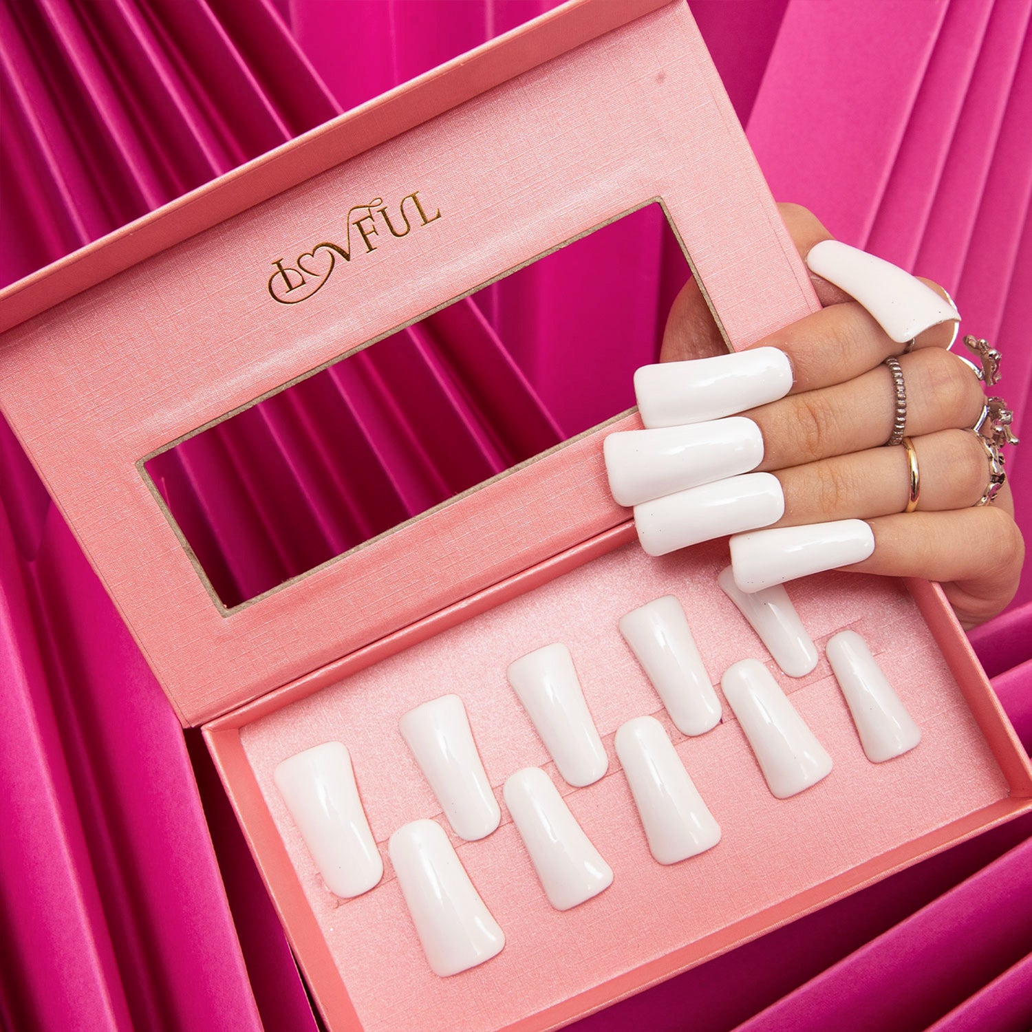 Long glossy white press-on nails displayed with an open pink Lovful box containing more white press-on nails against a pink fabric background.