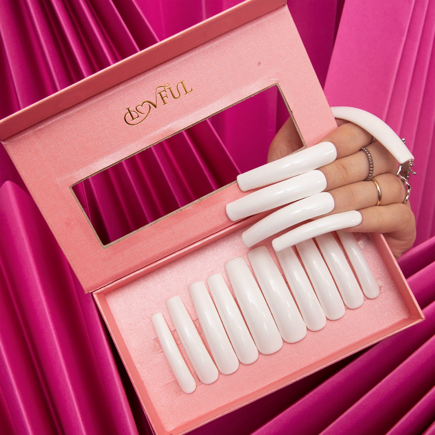 Lovful pink box showcasing white press-on acrylic nails set in various sizes, held by a hand with rings, with a pink fabric background.