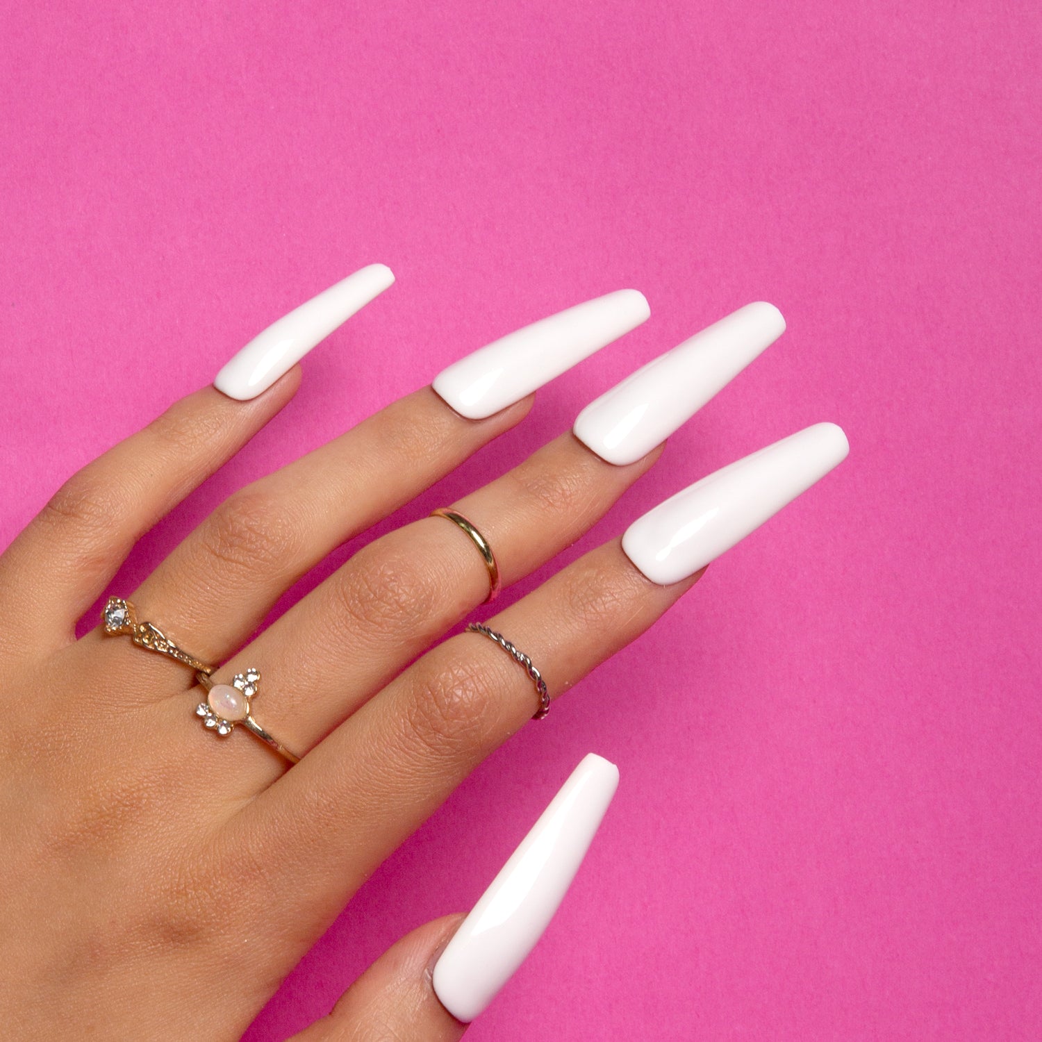 Hand with long, coffin-shaped, white press-on acrylic nails from Lovful against a pink background, decorated with multiple rings