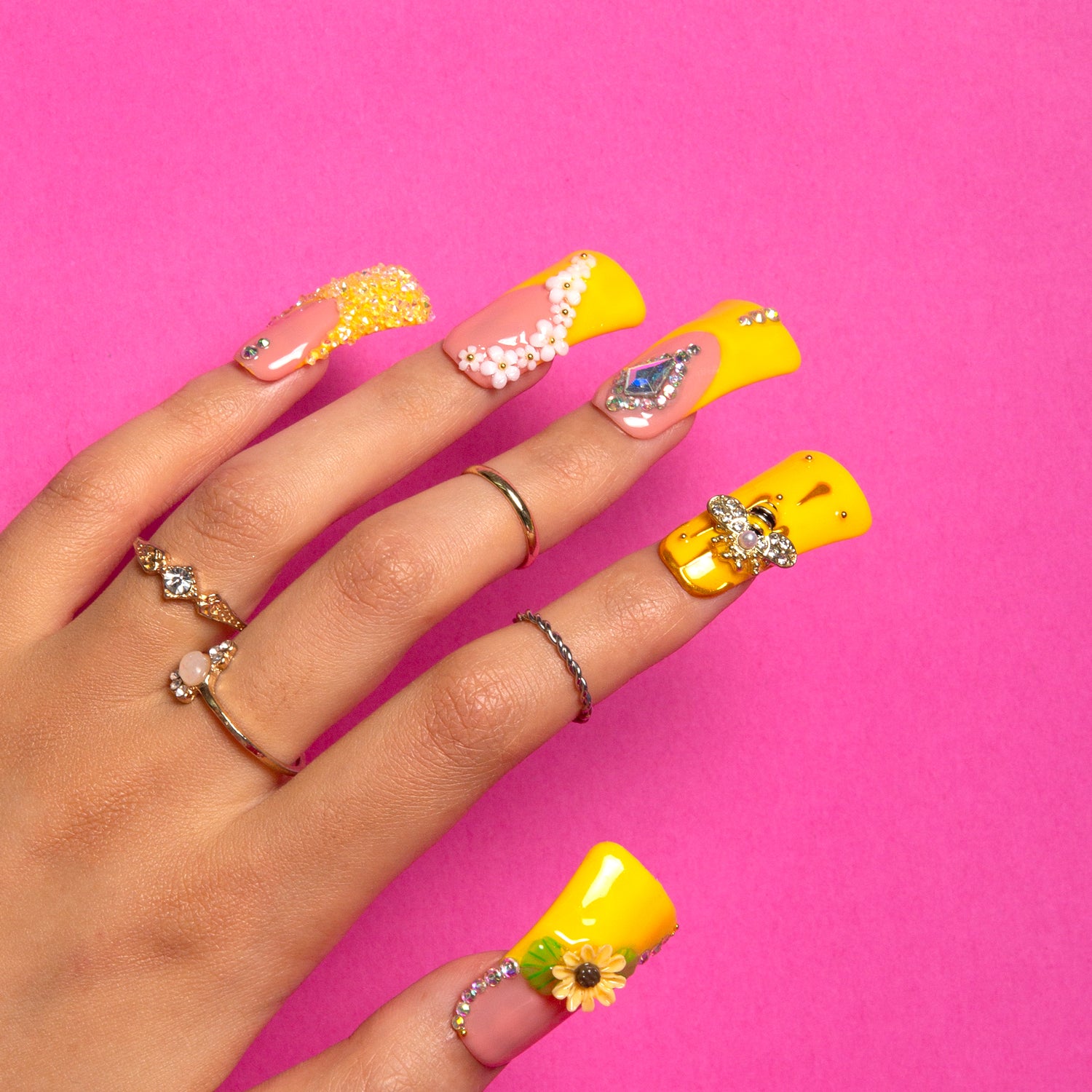 Bright yellow press-on acrylic nails with sunflower patterns, jewel embellishments, and bee accents on a pink background. The nails are modeled with gold rings adorned with stones.