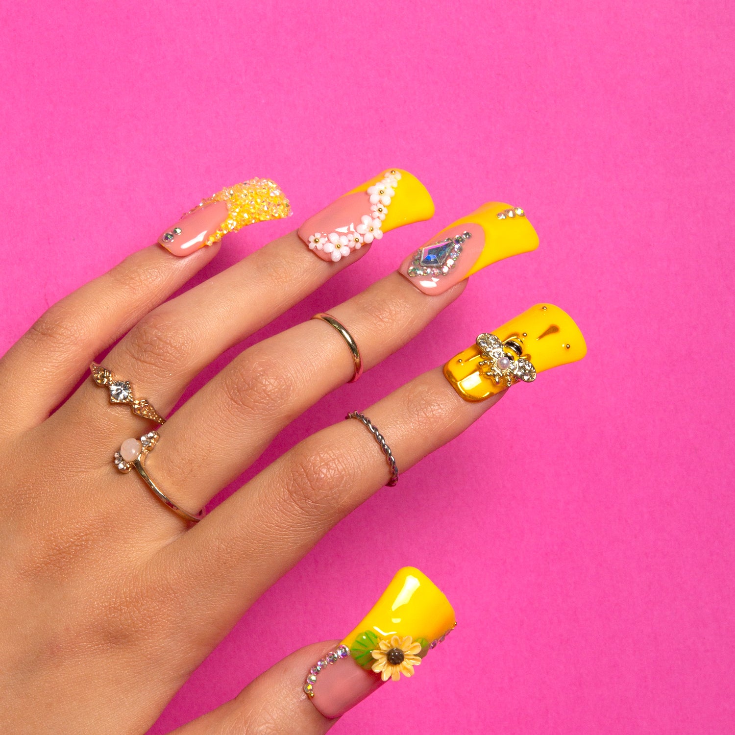 Hand with bright yellow press-on acrylic nails featuring sunflower patterns, bee accents, pearls, rhinestones, and intricate designs, complemented by multiple silver rings.