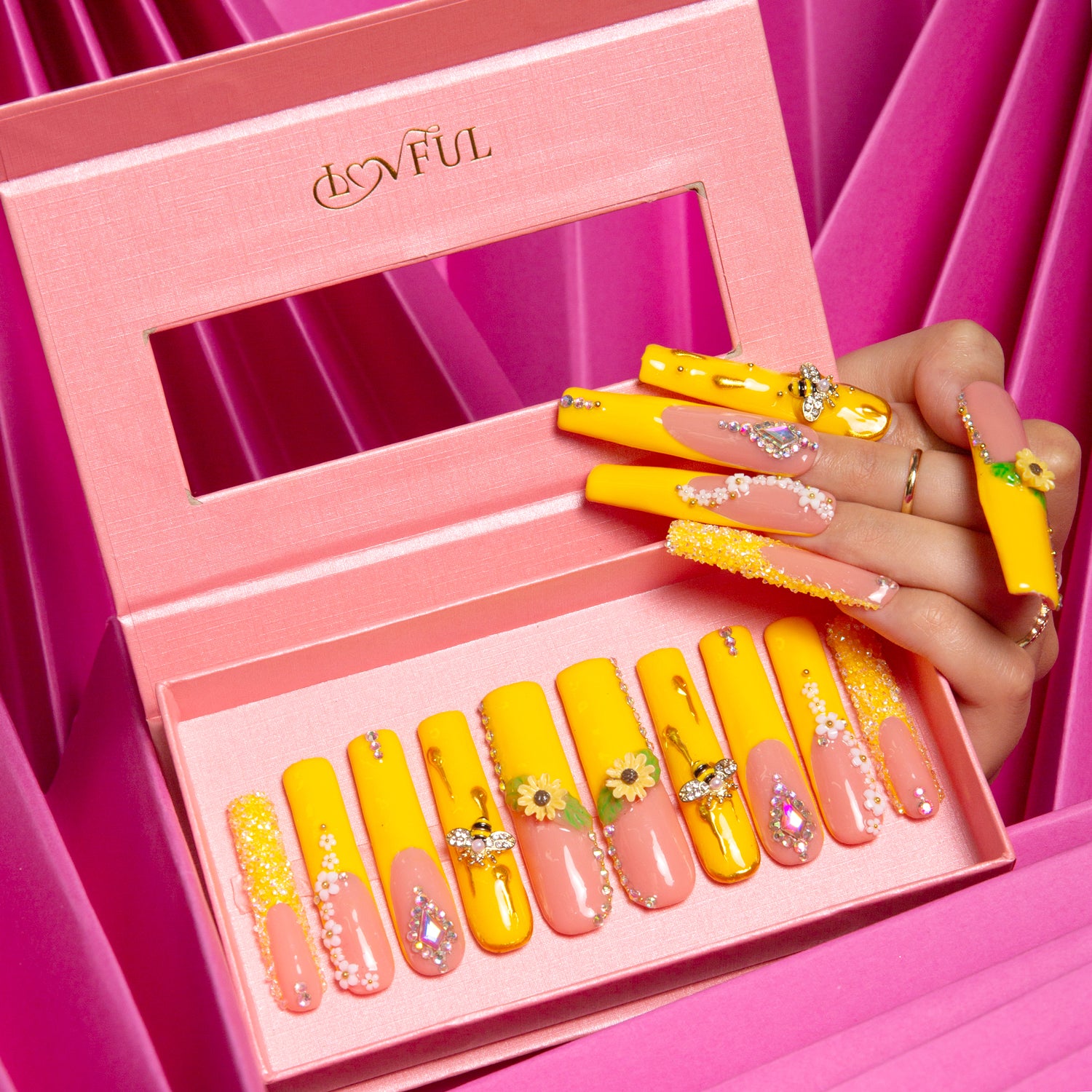 Yellow press-on acrylic nails with sunflower patterns and bee accents displayed in a pink Lovful box. Hand with matching nails holding the box.