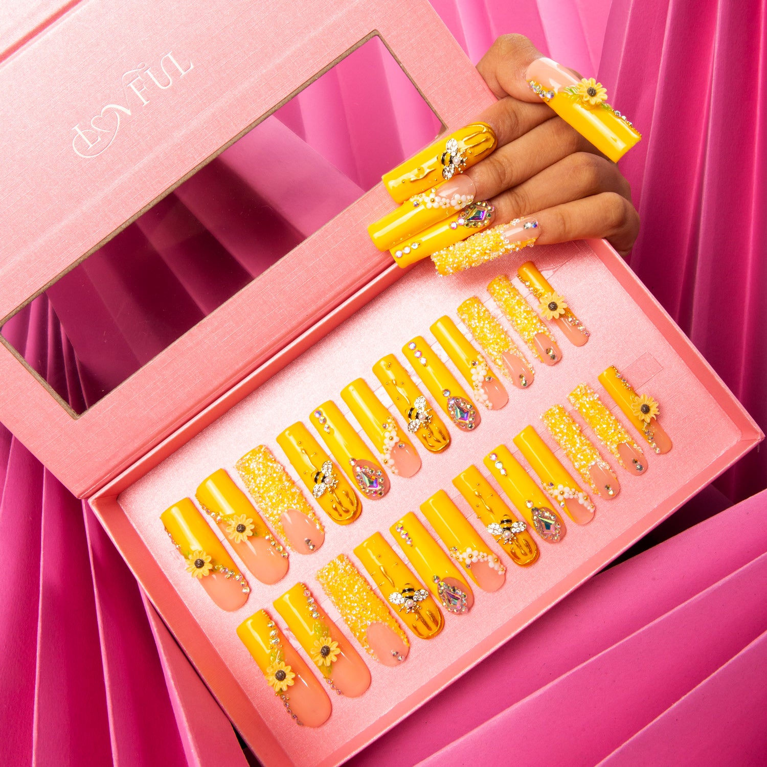 Bright yellow press-on nails with sunflower patterns and bee accents displayed in a pink box and on a hand model