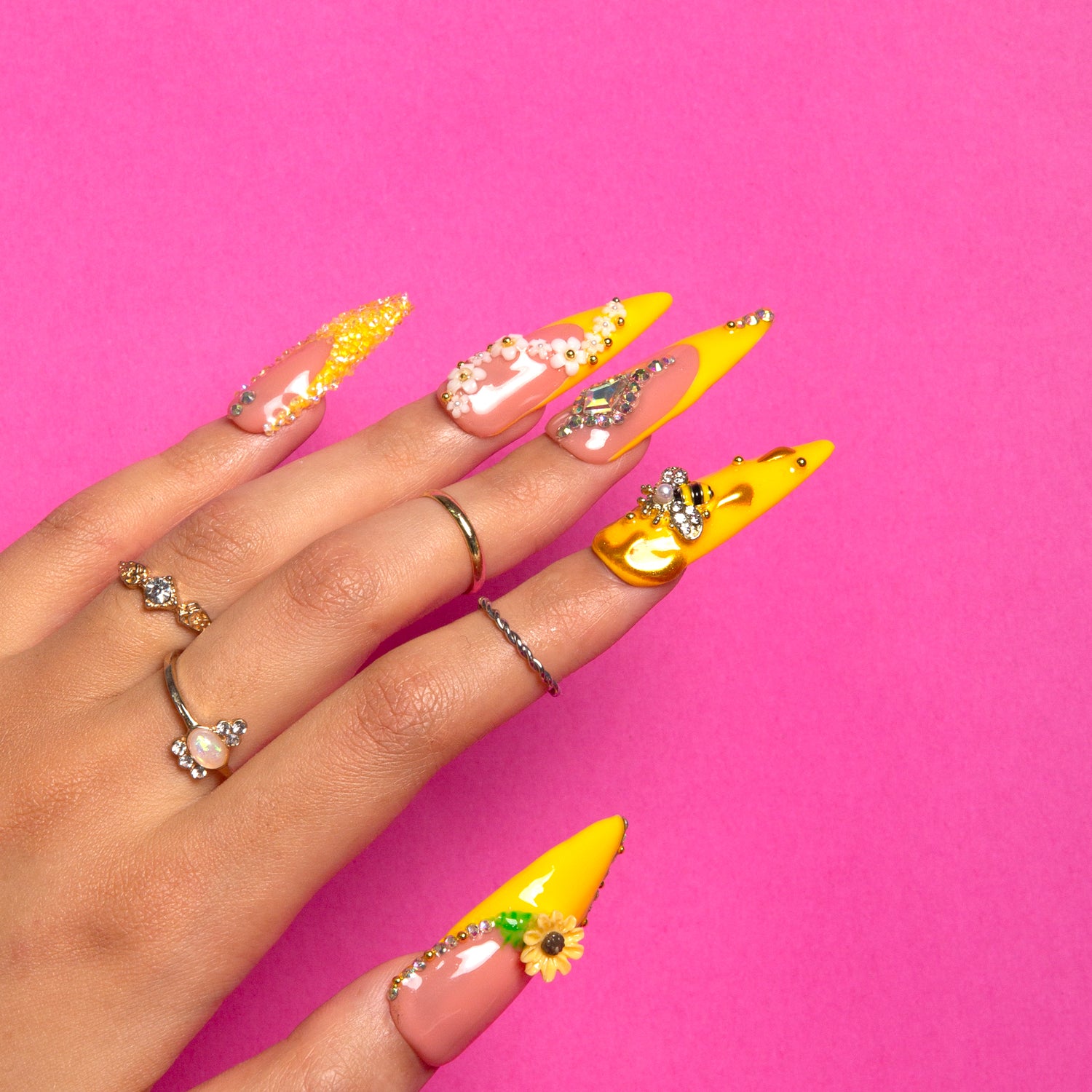 Hand showcasing long stiletto press-on nails with a bright yellow design, sunflower patterns, and bee accents, on a vibrant pink background, wearing multiple rings.