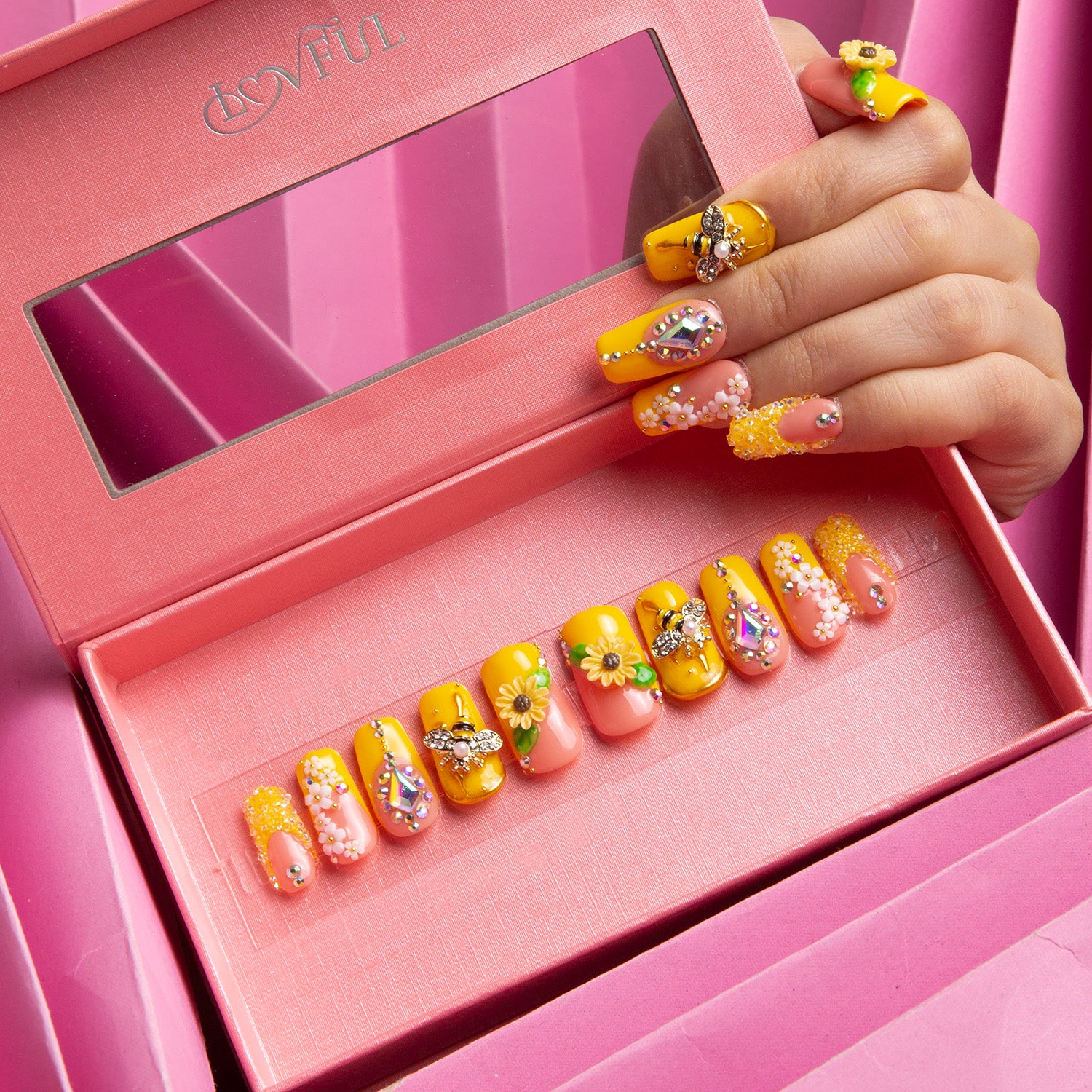 Hand displaying press-on acrylic nails with bright yellow sunflower patterns and bee accents inside a pink Lovful box, showcasing the 'Honey Bee - Square' design.