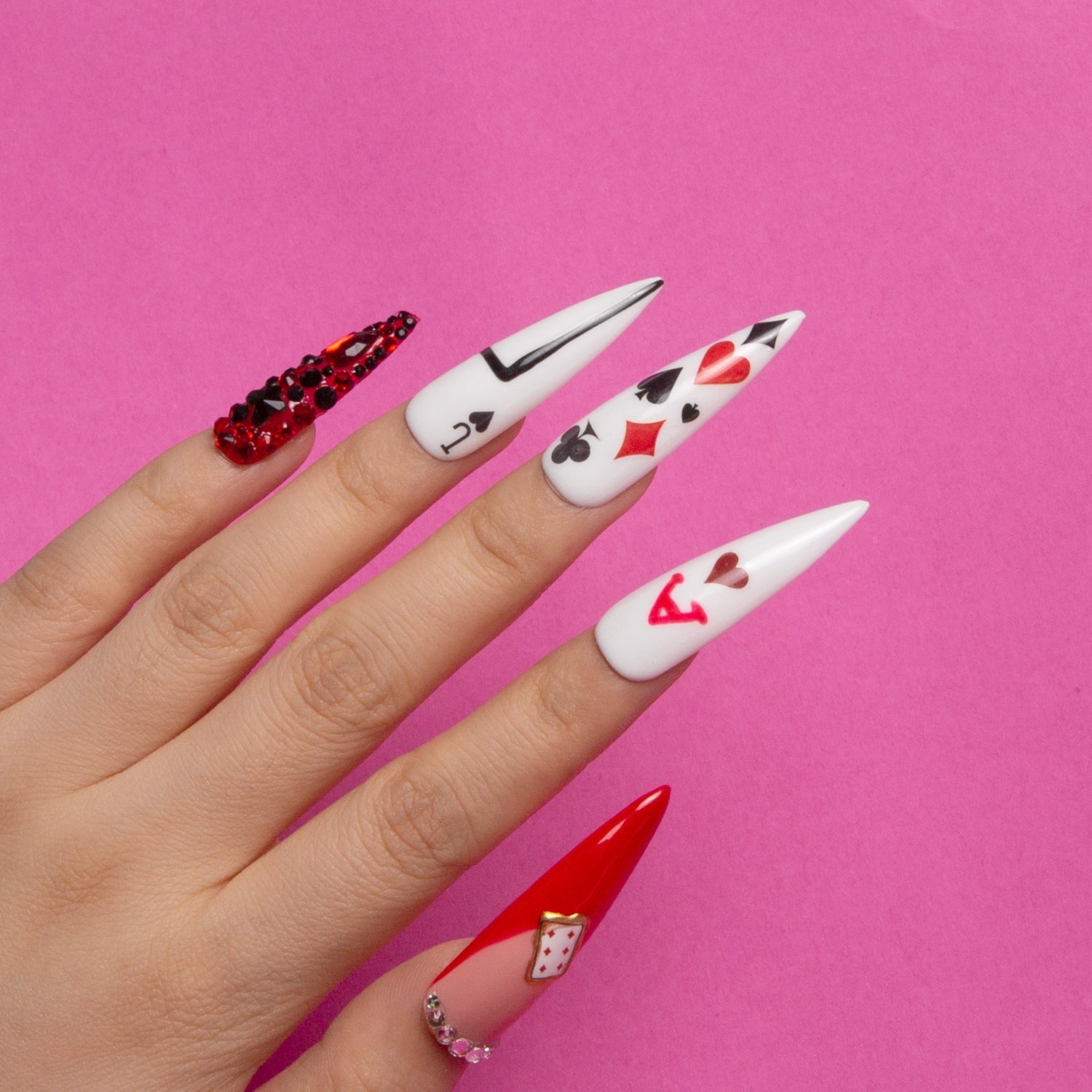 Hand with long stiletto-shaped press-on nails featuring playing card designs and rhinestones, inspired by the Queen of Hearts, against a pink background.