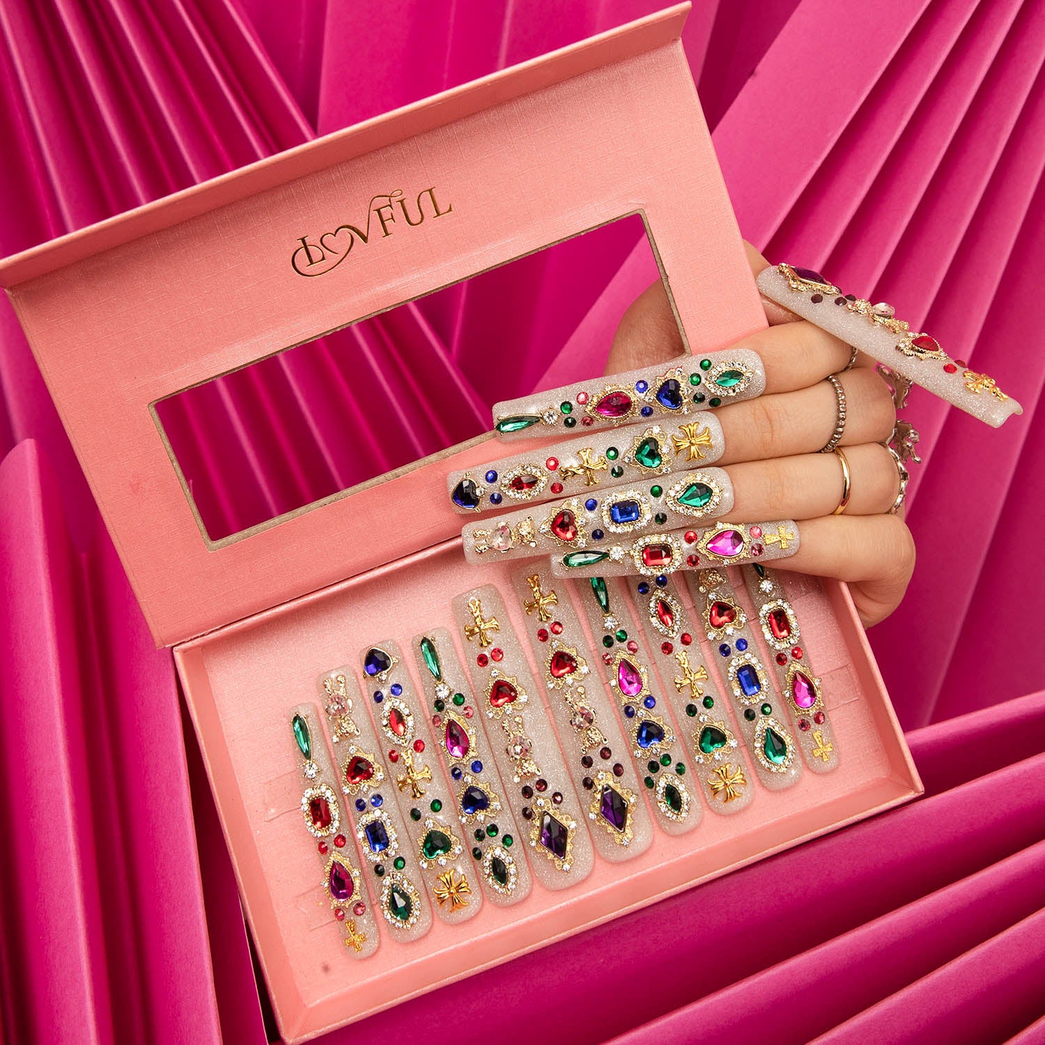 Hand showcasing H103 Treasure Trove square press-on nails by Lovful, decorated with colorful gems and glitter, set in a pink box against a pink pleated fabric background