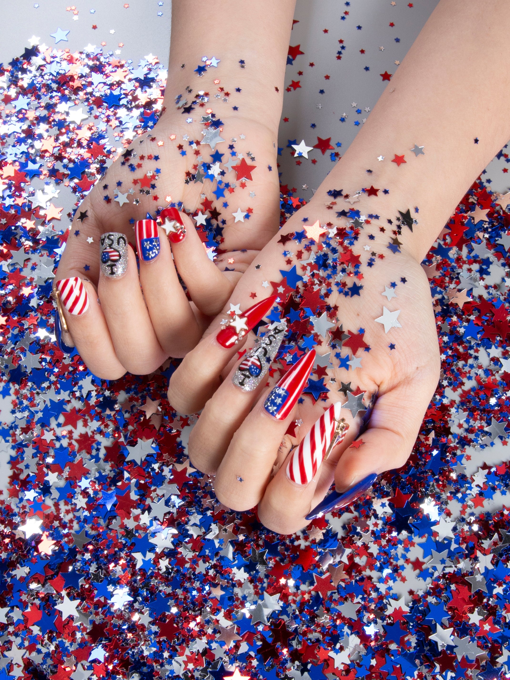 Hands adorned with American Dream press-on nails in red, white, and blue with stars and stripes designs, surrounded by red, white, and blue star-shaped confetti. Perfect for National Day celebrations and July 4th parties.