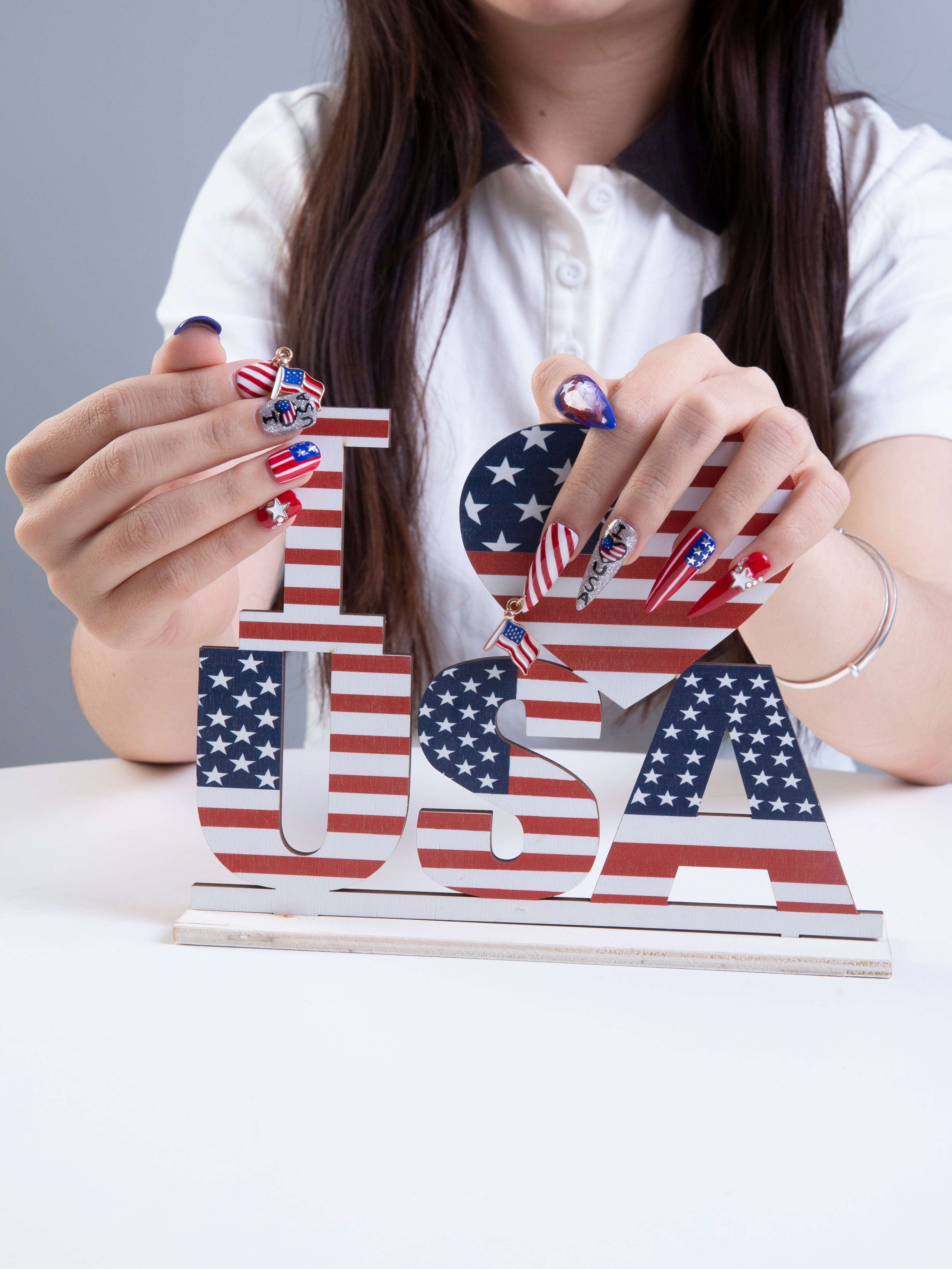 Person with American flag-themed press-on nails holding 'I a??¡§¡§ USA' decor. Patriotic red, white, and blue nail design perfect for Independence Day celebrations and July 4th parties.