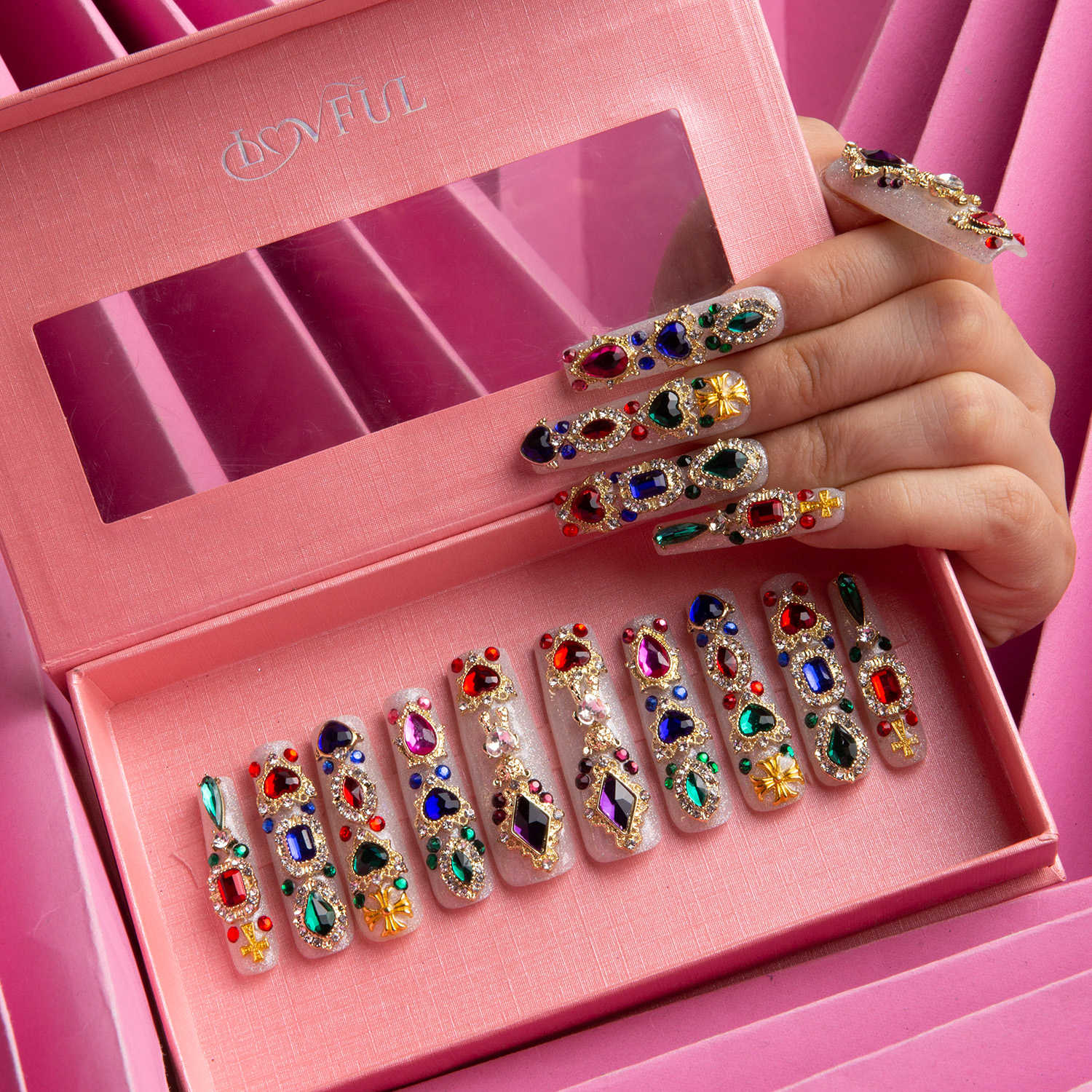 Hand wearing press-on nails adorned with colorful gems and glitter above a pink box containing a set of similar detailed and vibrant press-on nails.