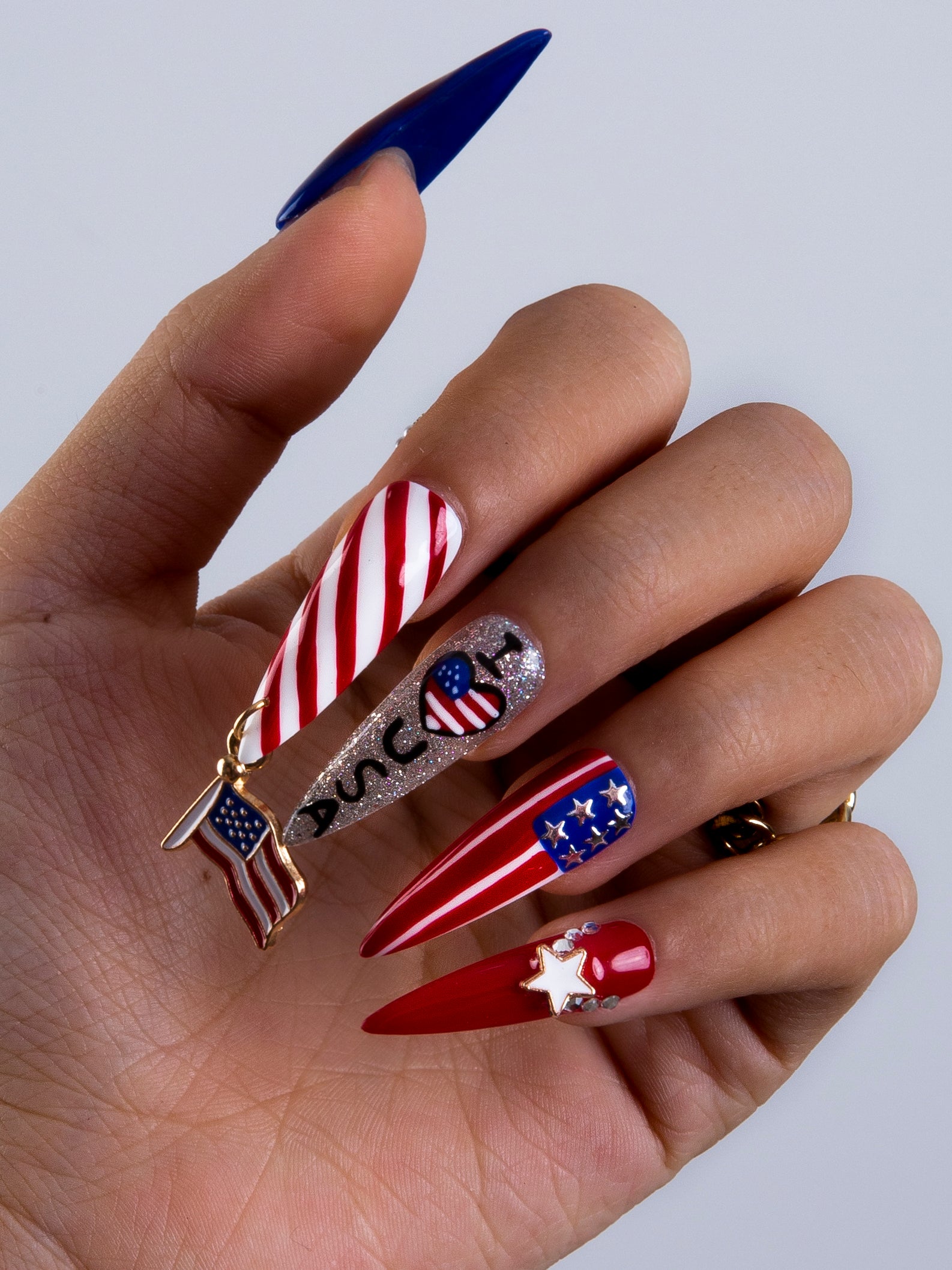 Hand with five press-on nails from the 'American Dream' collection by Lovful. The designs include red and white stripes, glittery silver with flag motifs and 'LOVE' text, solid red with a white star, blue with white stars, and solid blue. Ideal for National Day and July 4th celebrations.