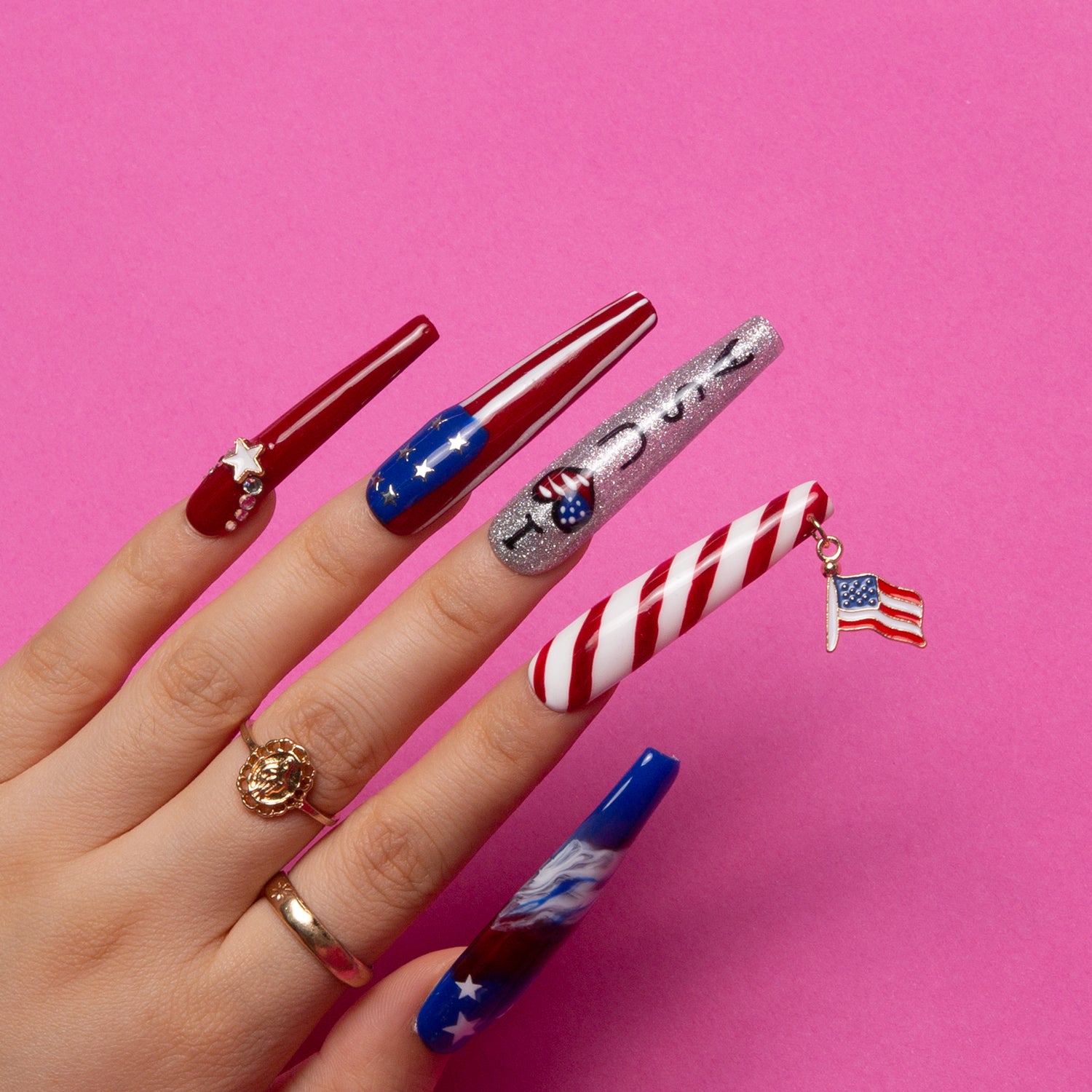 Hand displaying 'American Dream' press-on nails with patriotic designs including red, white, and blue, stars, stripes, and mini American flag charm on a pink background.