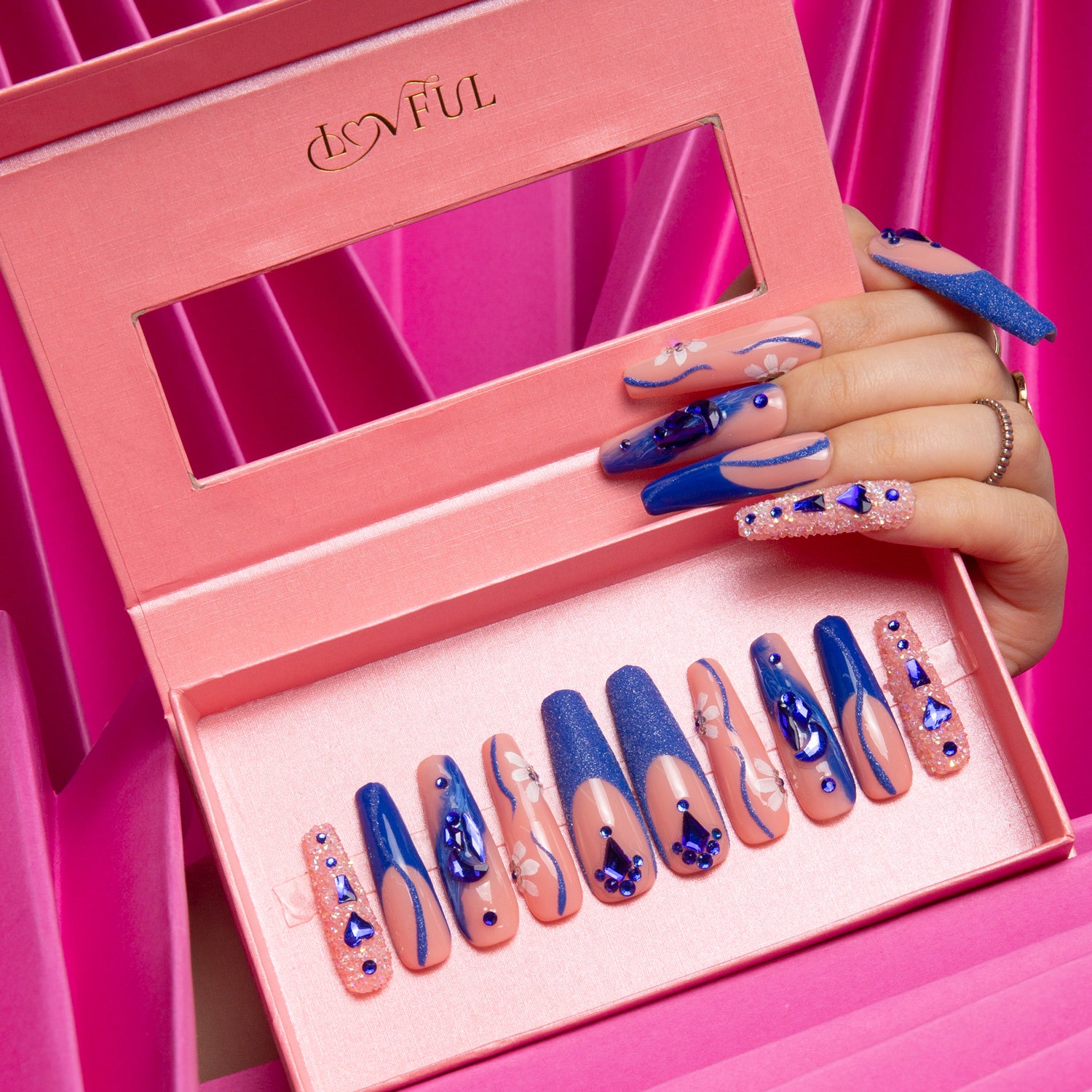 Hand showcasing blue suede press-on nails by Lovful.com, holding a pink box with a set of blue press-on nails featuring blue French tips, curvy lines, crystal decorations, and blue heart-shaped gems. Pink draped fabric in the background.