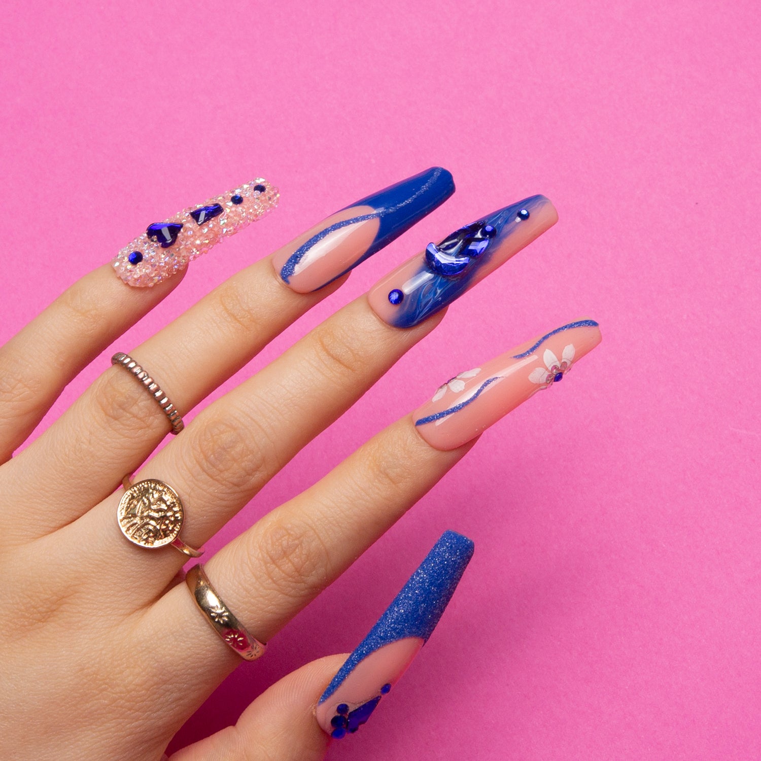 Hand showcasing Blue Suede coffin-shaped press-on nails with blue French tips, curvy lines, crystals, heart-shaped gems, sparkles, and flower designs against a pink background.