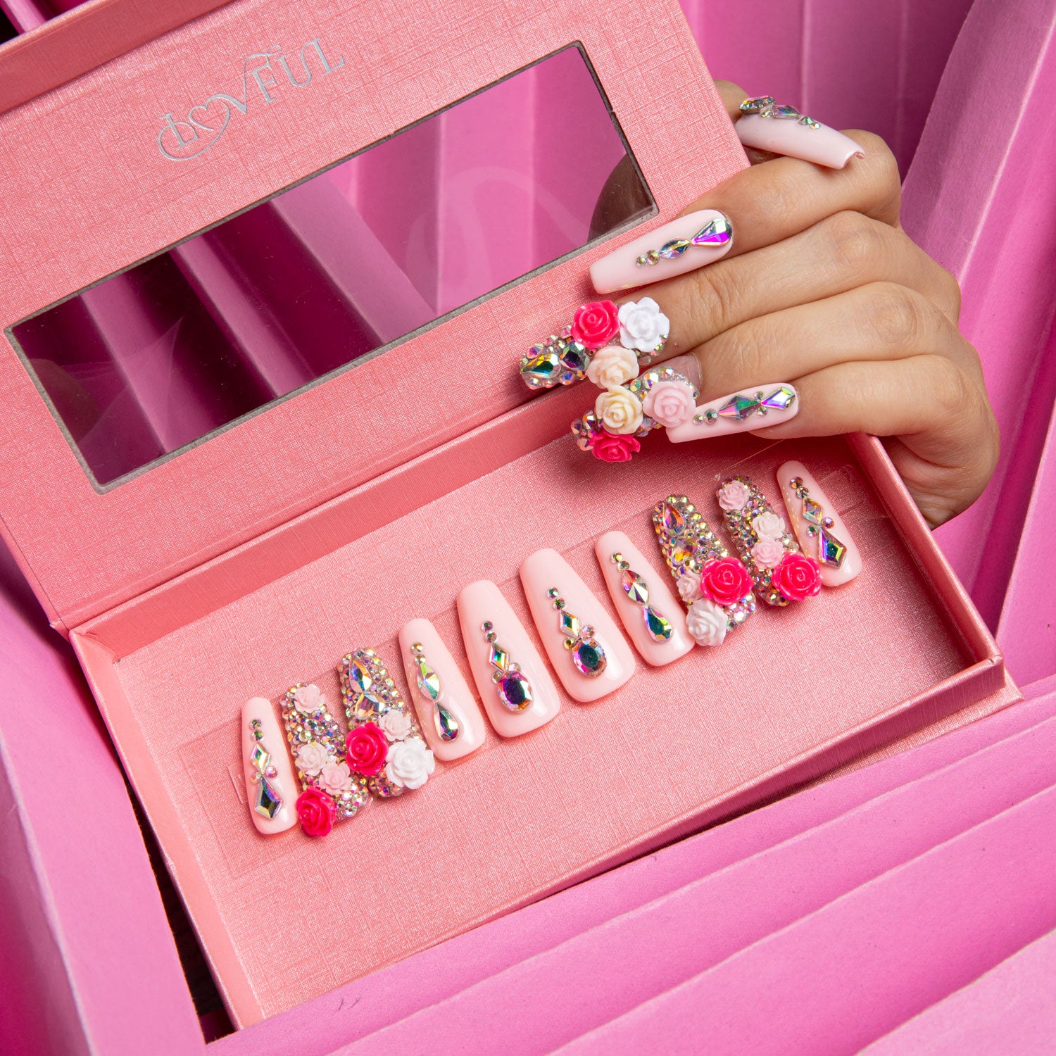 Hand with rose garden-themed press-on nails detailed with rhinestones and floral designs above an open pink Lovful box displaying matching press-on nails, all set against a pink box background.