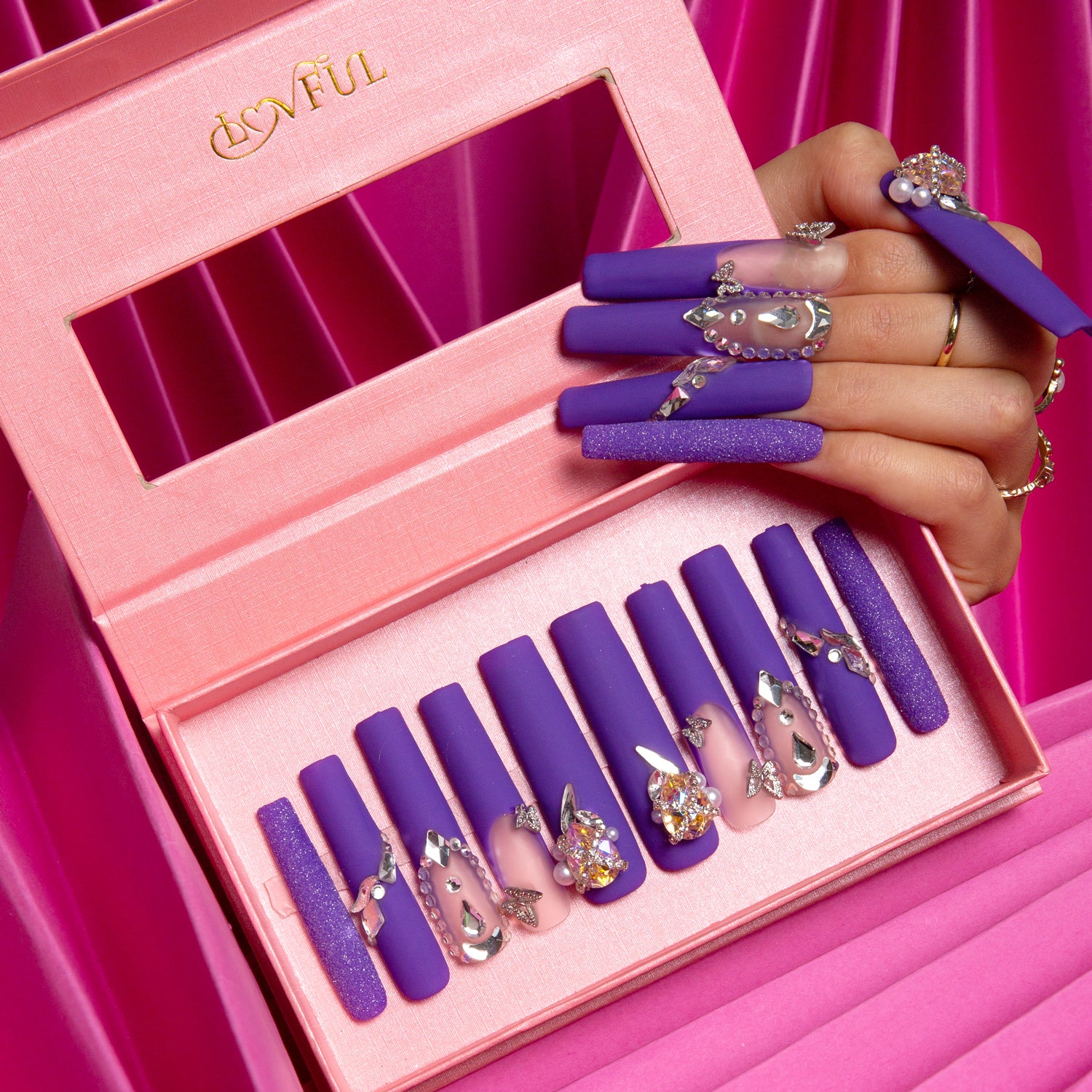 Mystique Veil press-on nails by Lovful displayed in a pink box. The deep purple nails feature butterfly designs, gem accents, and glitter overlays. A hand with the nails applied holds the box, set against a pink fabric background.