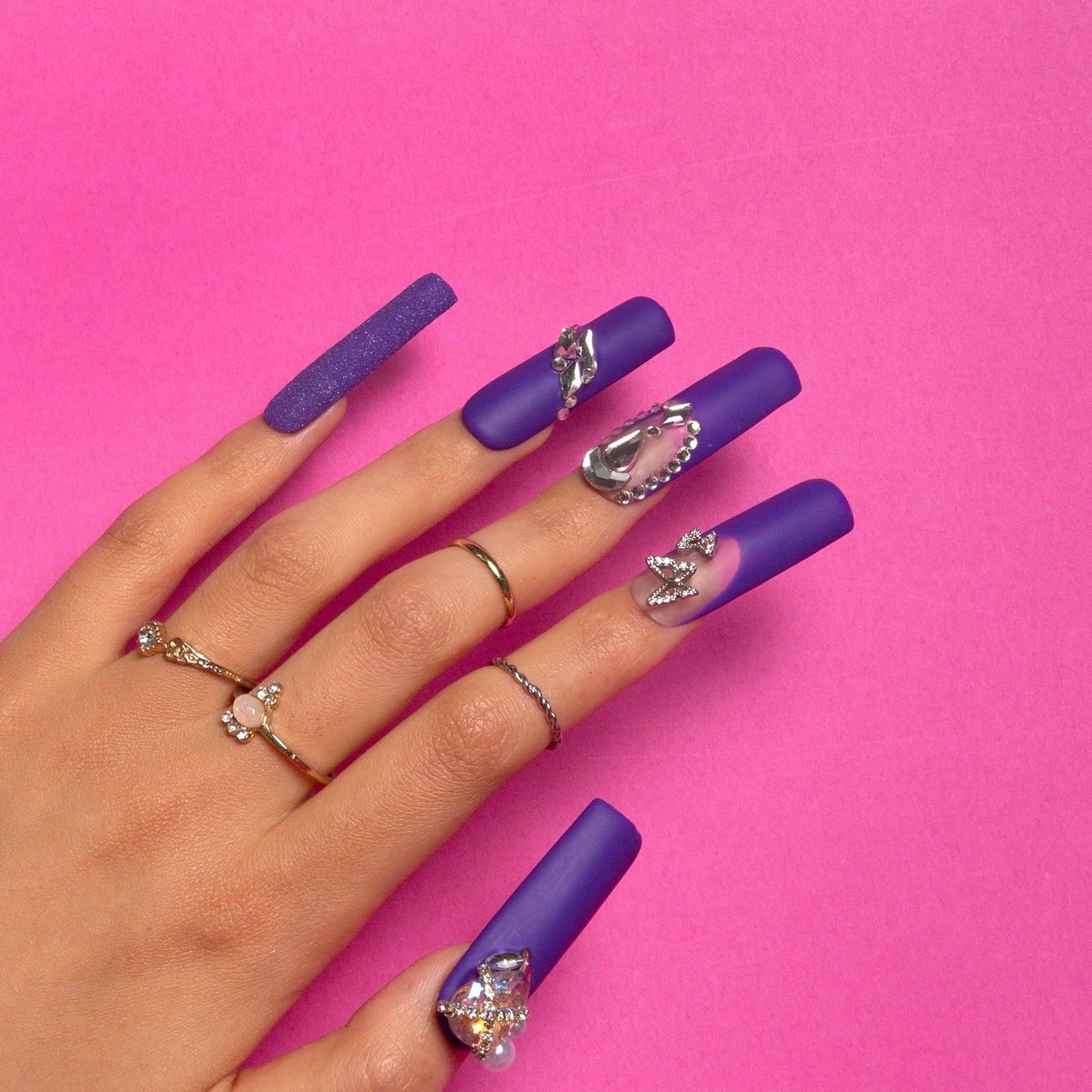 Hand showcasing Mystique Veil, square-shaped deep purple press-on nails with butterfly decorations, gem accents, and a shimmering glitter overlay, against a pink background.