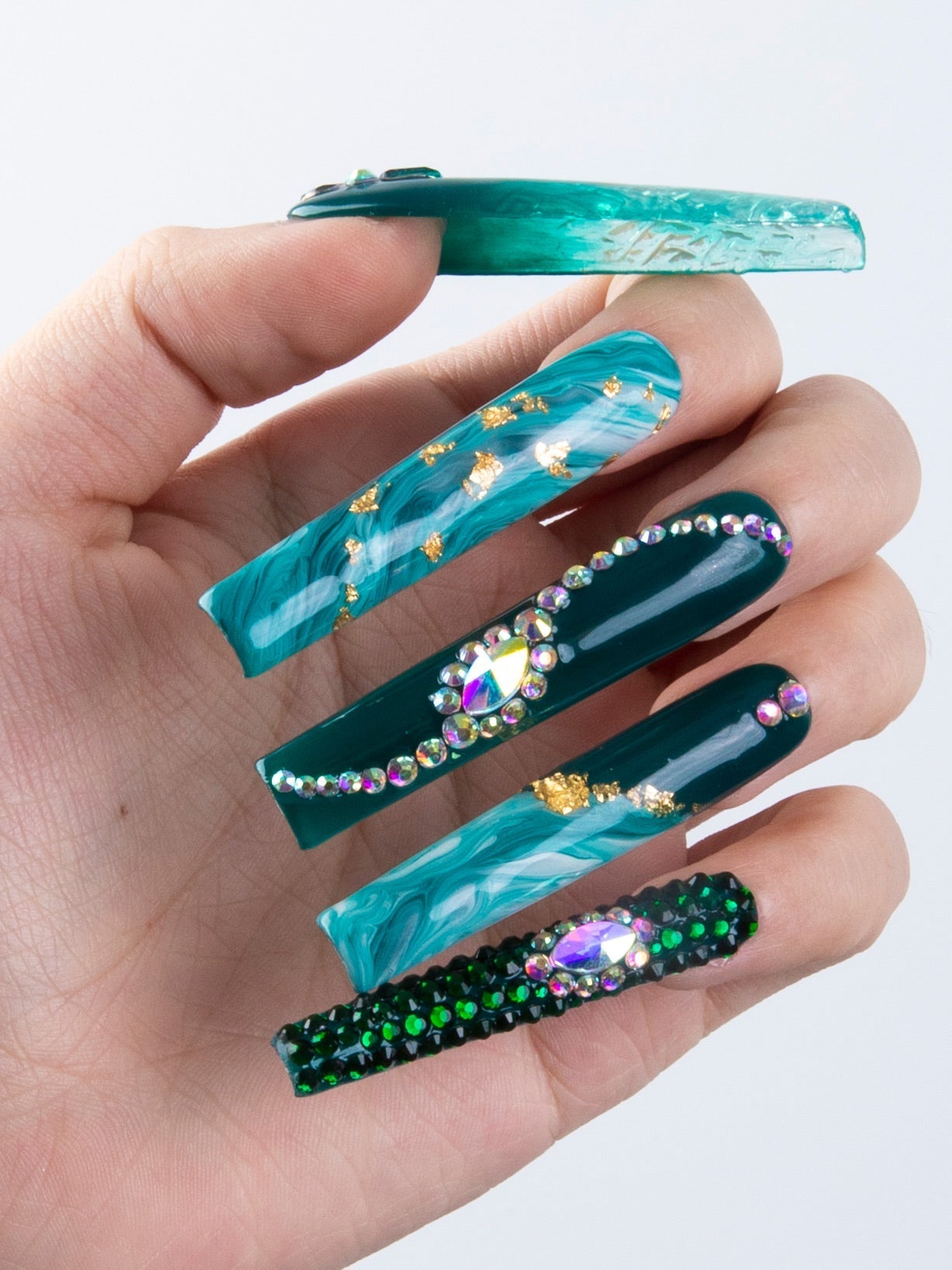 Hand with five Emerald Envy press-on nails featuring green liquid flowing design, gold decor, and rhinestones capturing the essence of woodland allure.