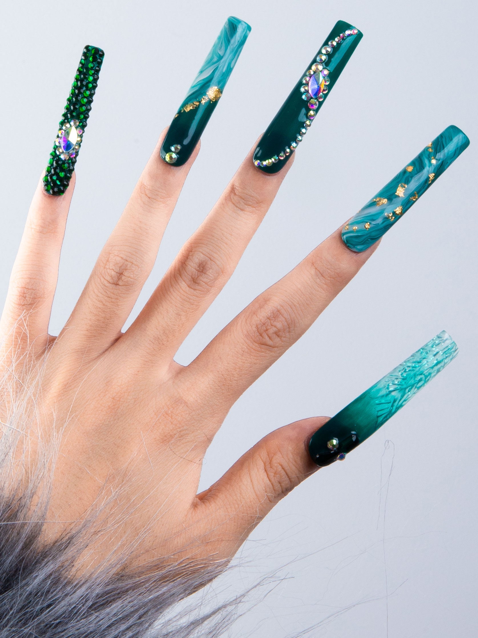 Hand showcasing Lovful.com's Emerald Envy press-on acrylic nails with intricate gold decor, rhinestones, and green liquid flow designs evoking lush foliage.