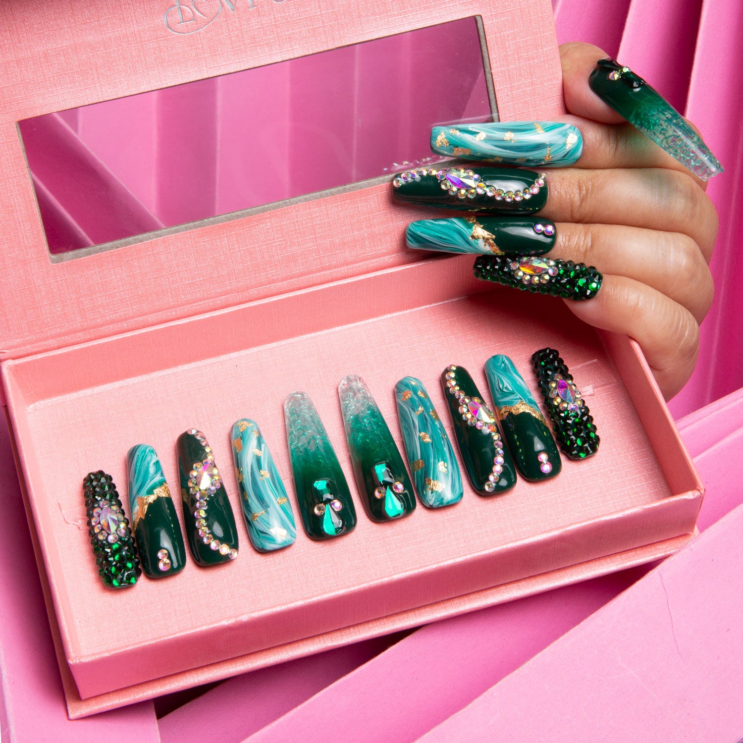 Lovful Emerald Envy press-on nails set in a pink box. Designs feature gradient green shades, gold decor, and a mesmerizing liquid flowing effect. A hand with applied nails is holding the box.