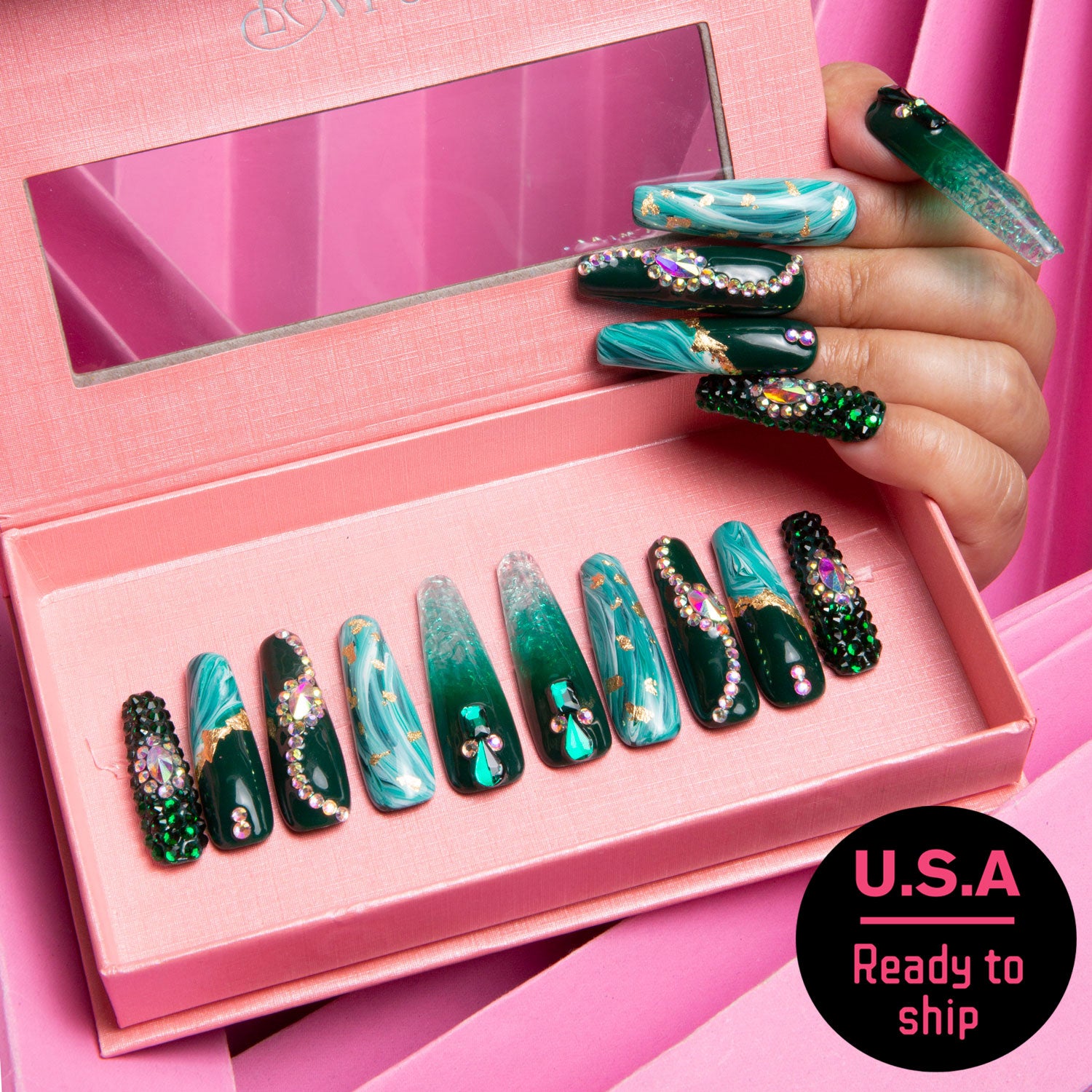 Hand showcasing long, emerald green and gold press-on nails, holding a pink box of Emerald Envy Coffin-shaped nails. Text reads 'U.S.A Ready to ship.'