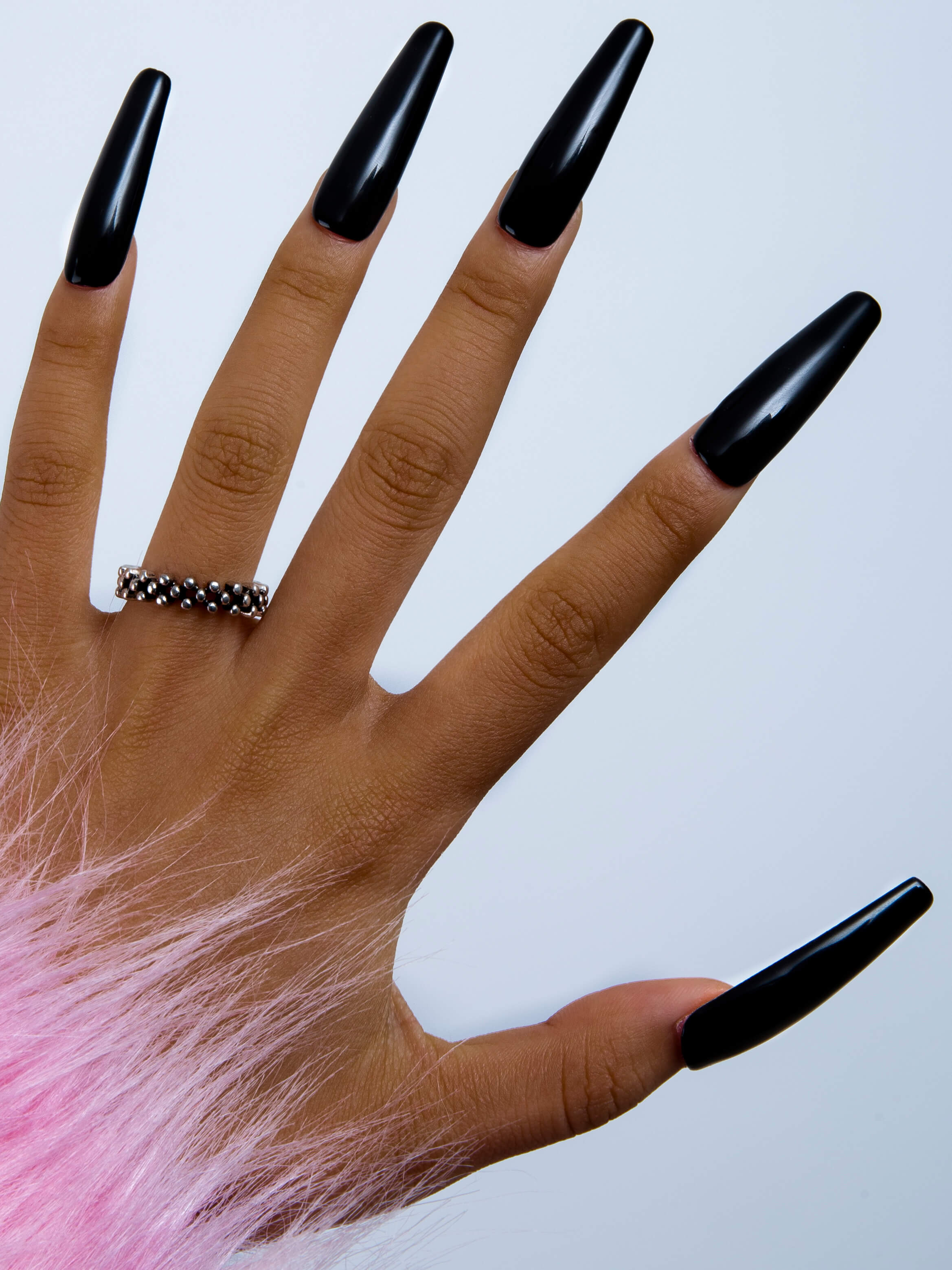 Hand with long black glossy press-on nails from Lovful's H11 Dark Angel collection, showcasing sleek and shiny finish with a beaded silver ring and pink fuzzy wrist accent