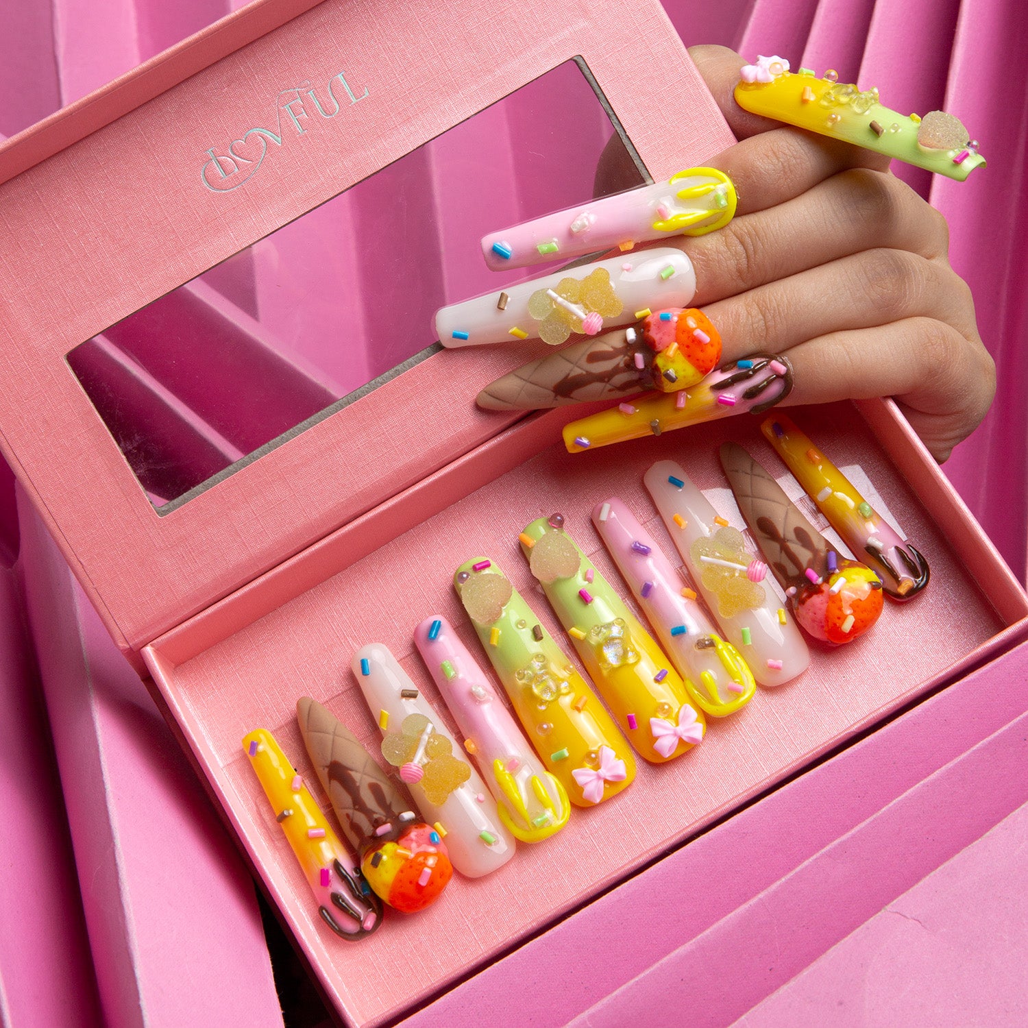 Set of 'Ice Queam - Coffin' press-on nails by Lovful featuring colorful 3D decorations of sprinkles, candy canes, mini ice cream cones, and other playful elements in a pink box with a mirrored lid. A hand is showcasing some of the nails.