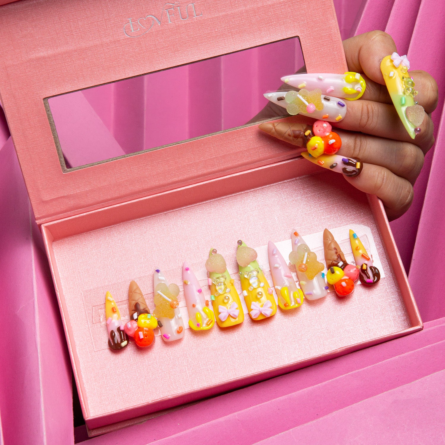 Set of press-on stiletto nails in 3D designs featuring sprinkles, candy canes, ice cream cones, and cherries inside a pink display box. A hand holding one nail with similar sweet-themed decorations.