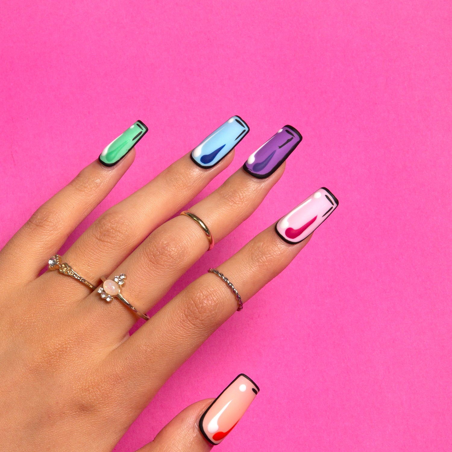 Colorful Pop press-on nails in square shape with green, light blue, purple, pink, and peach pop art designs against a pink background. The hand is adorned with gold and silver rings.