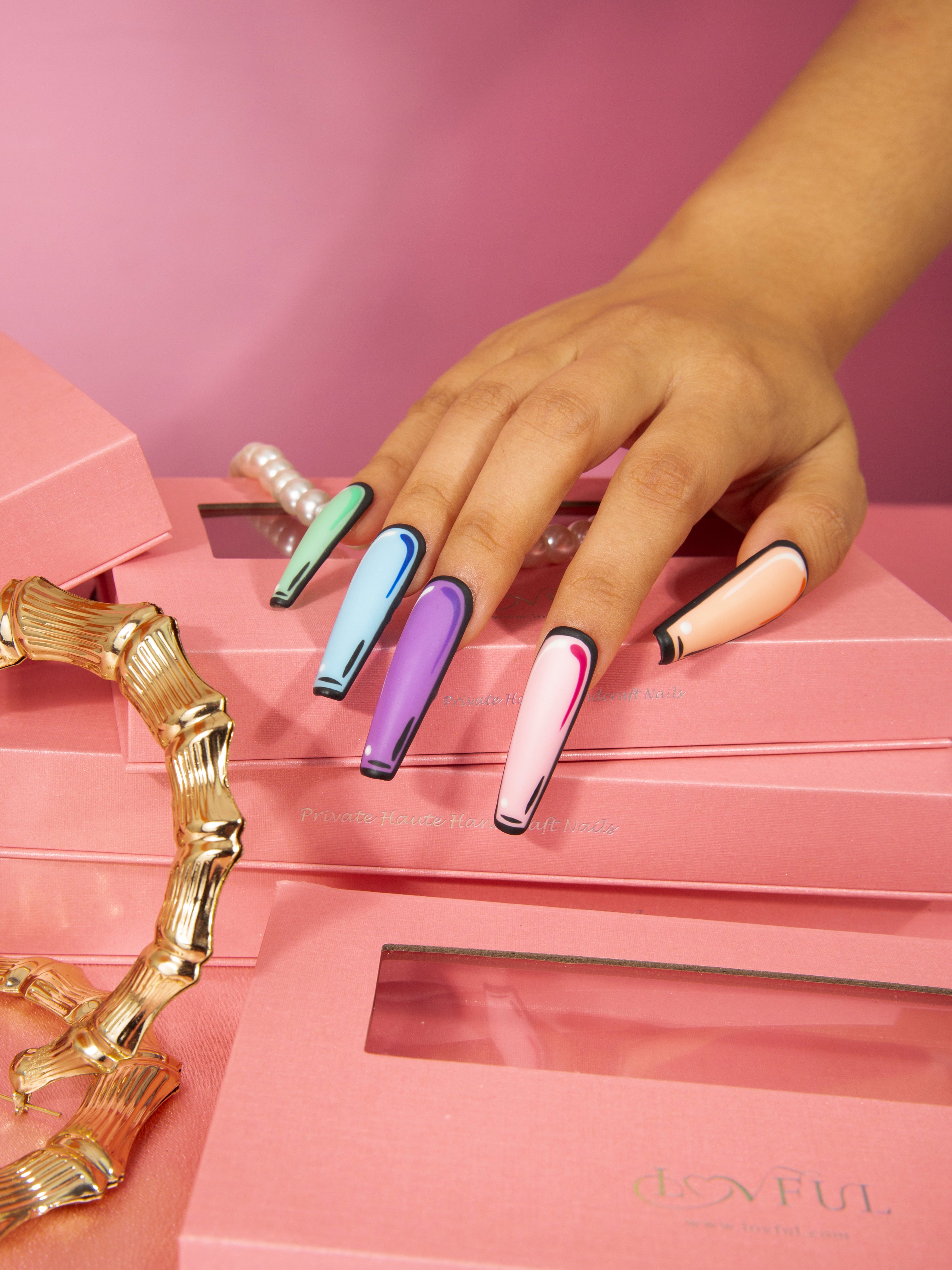 Hand with vibrant pop art press-on nails in blue, purple, pink, and peach colors against pink Lovful product boxes. Gold jewelry enhances the stylish presentation.