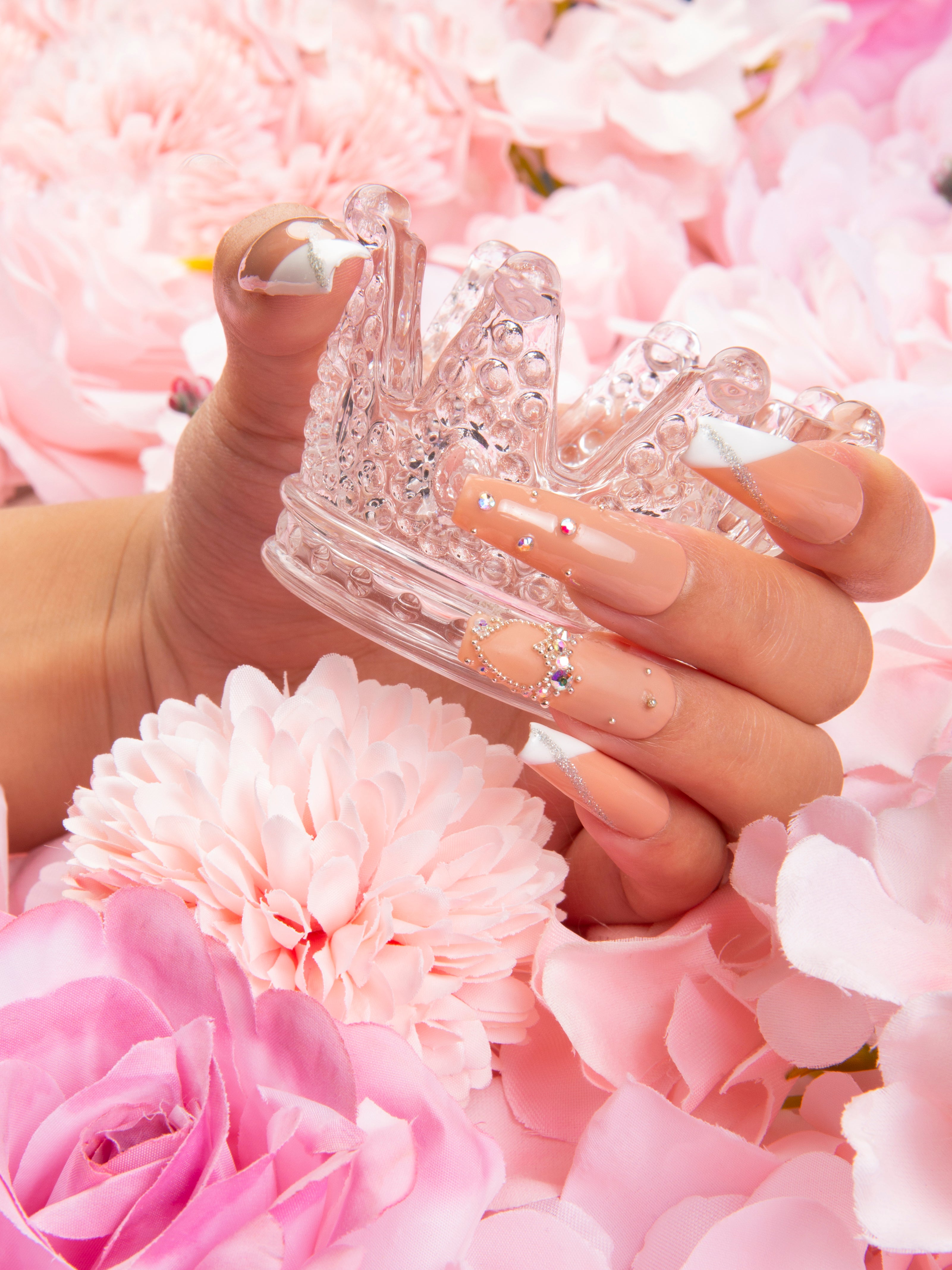 Hand with 'Lovely Kitten Heels' press-on acrylic nails in French tip design with glitter and rhinestones on a clean white base holding a clear crystal crown, surrounded by pink and white flowers.