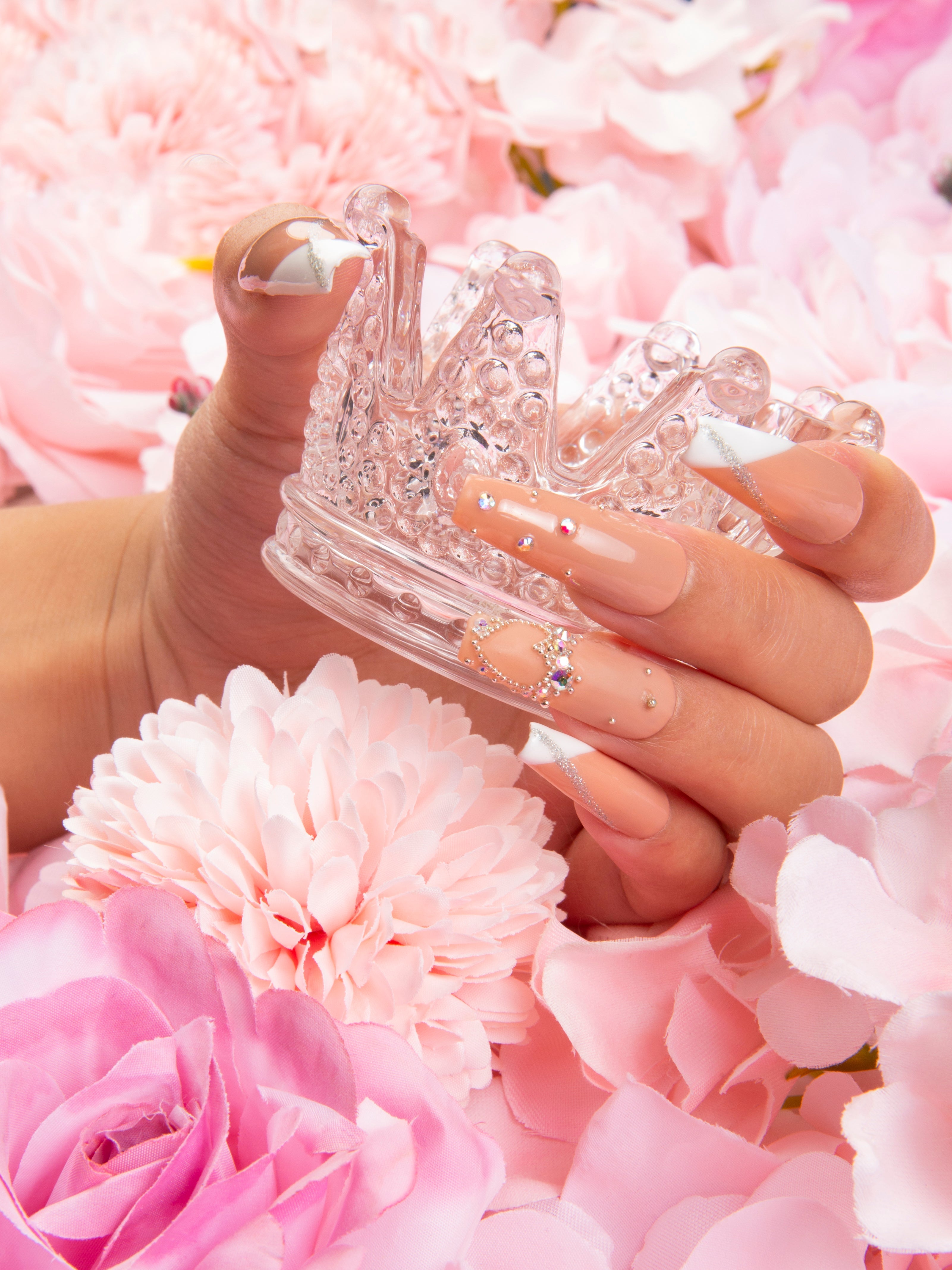 Hand displaying 'Lovely Kitten Heels' press-on nails with French tips and glitter accents, holding a clear acrylic crown amidst pink flowers. Lovful press-on acrylic nails.