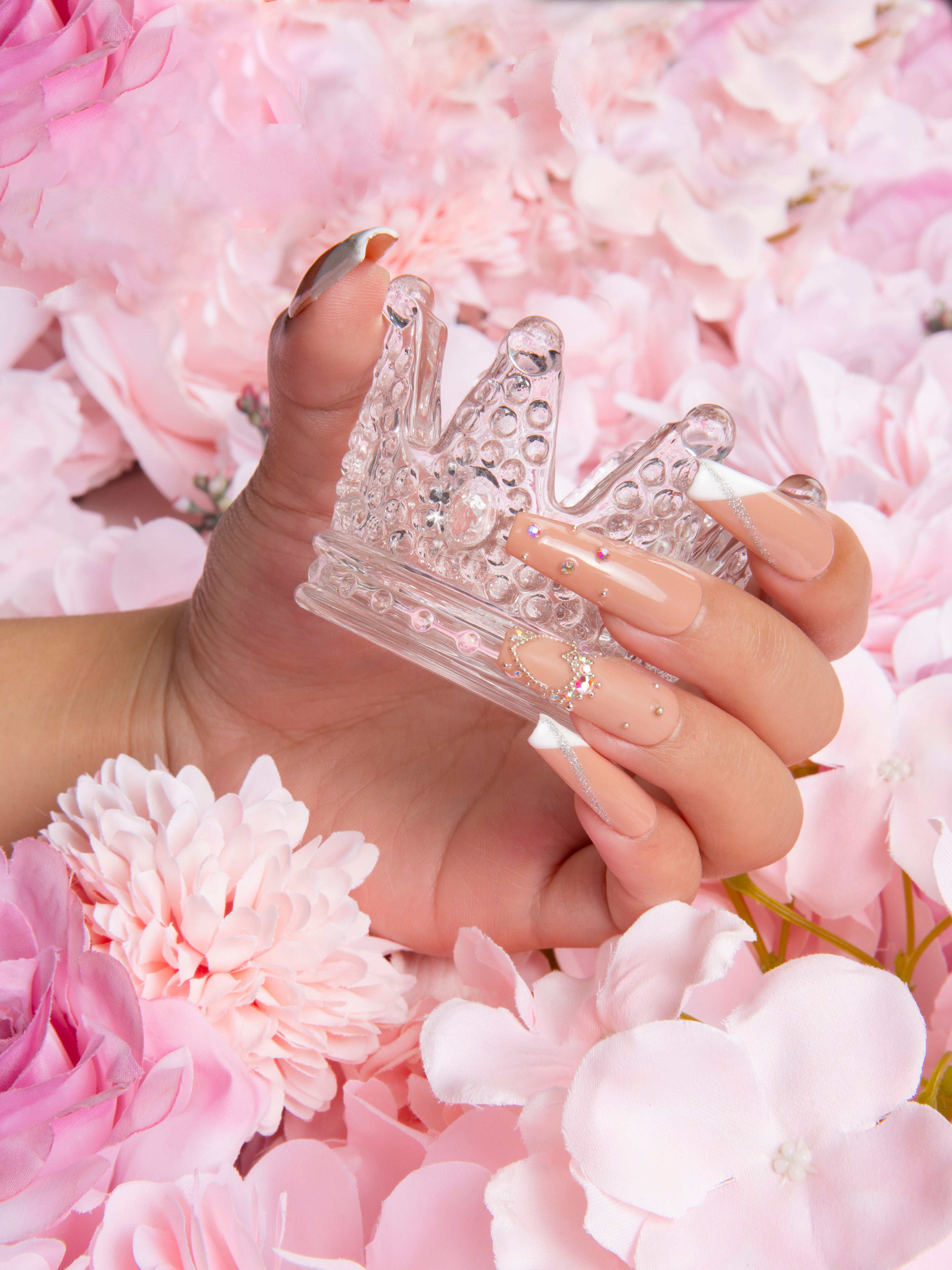 Hand with 'Lovely Kitten Heels' press-on nails featuring classic French tips and subtle glitter holding a clear crown figurine, surrounded by pink flowers.