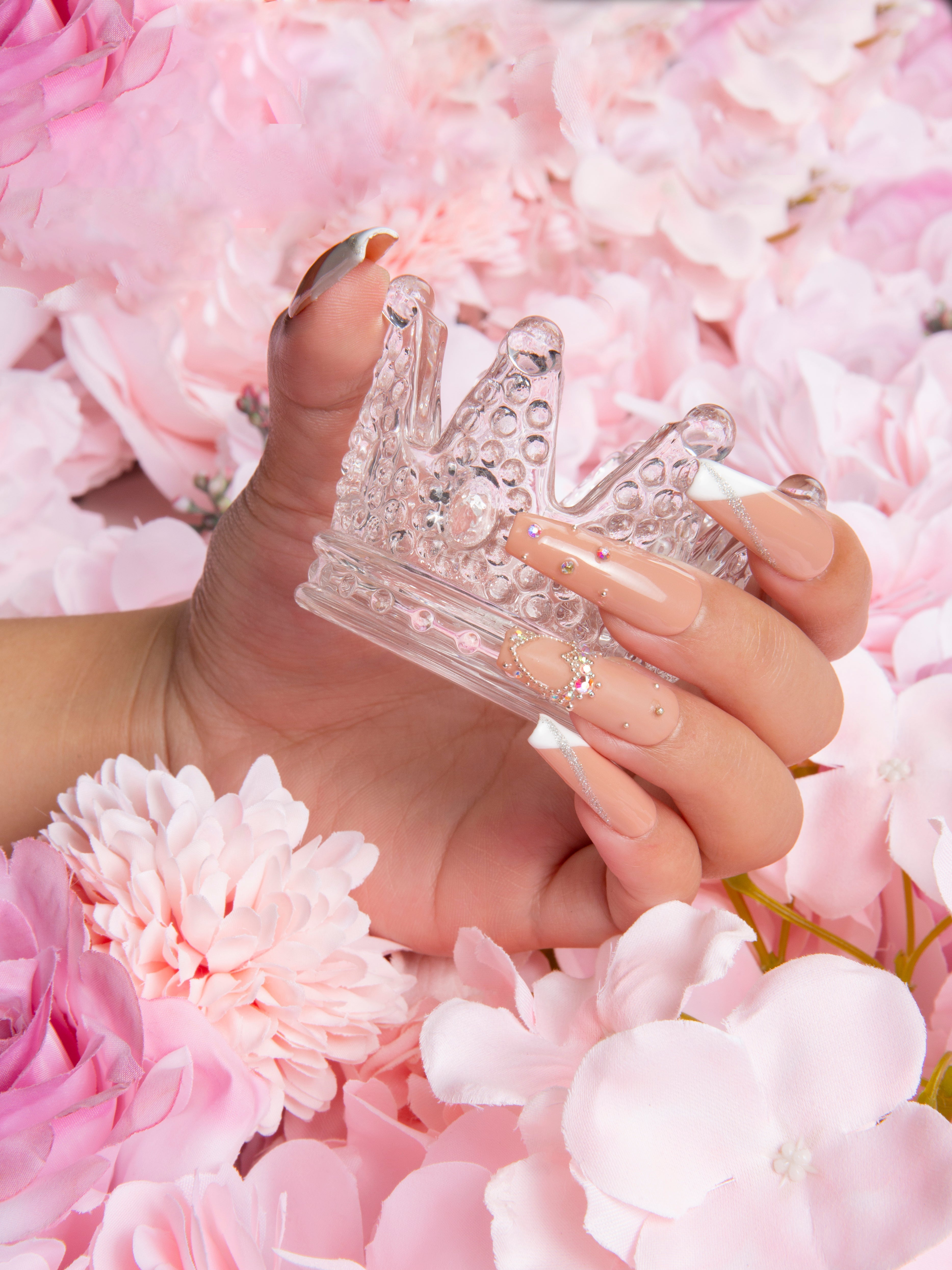 Hand with press-on acrylic nails featuring French tips, glitter, and embellishments, holding a translucent crown among light pink and white flowers.