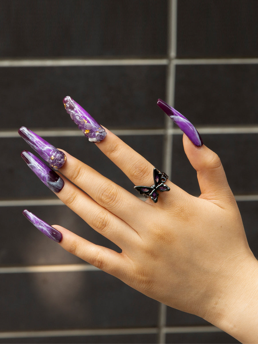 Hand with Dream Amethyst press-on nails featuring purple designs and hand-drawn patterns. The person is wearing a black butterfly ring, against a backdrop of dark tiles.