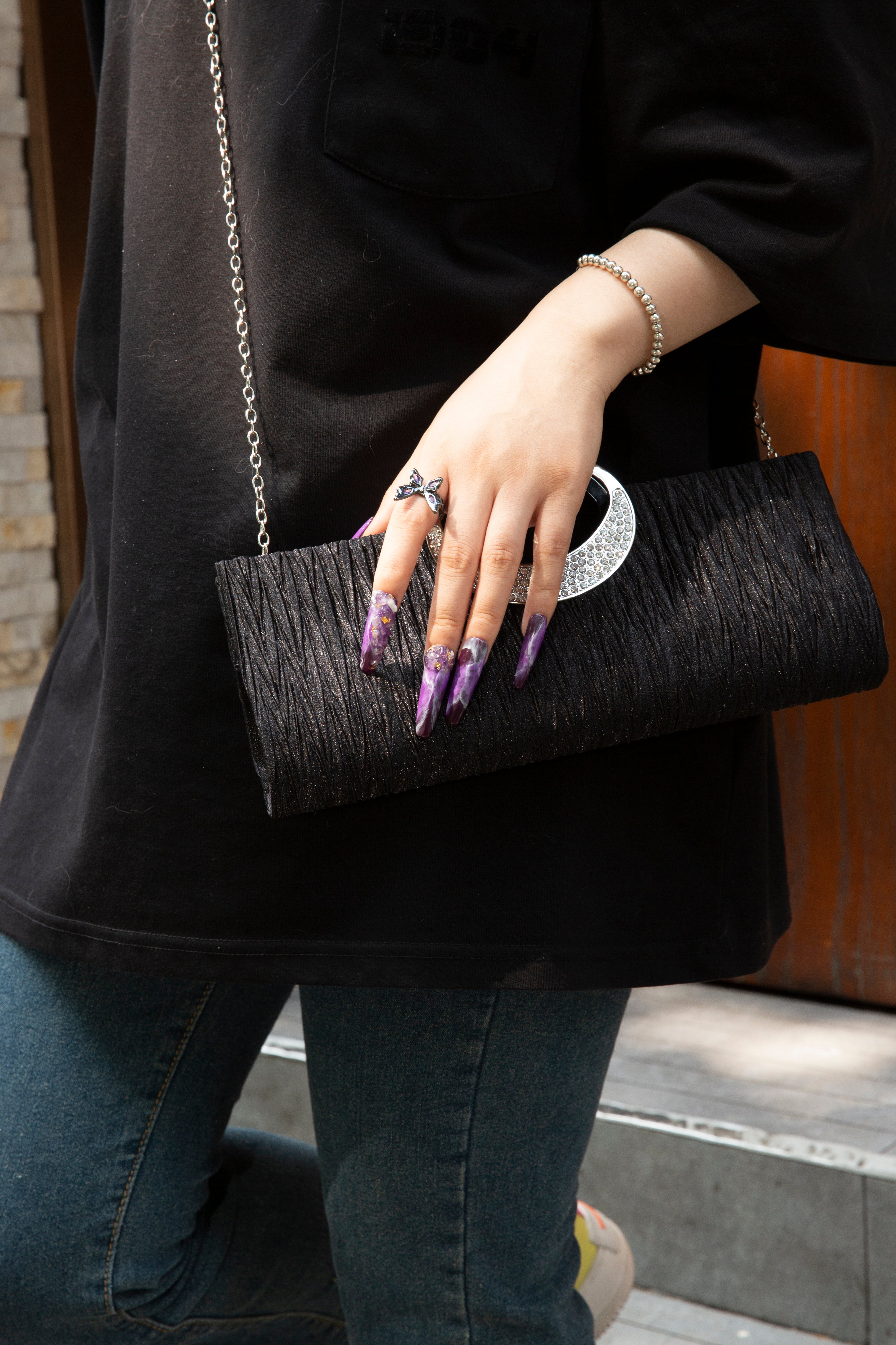 Person holding a black textured clutch with a circular silver accent, showcasing intricate purple press-on nails with mystical designs, wearing a black shirt, blue jeans, a bracelet, and a silver ring with a large stone