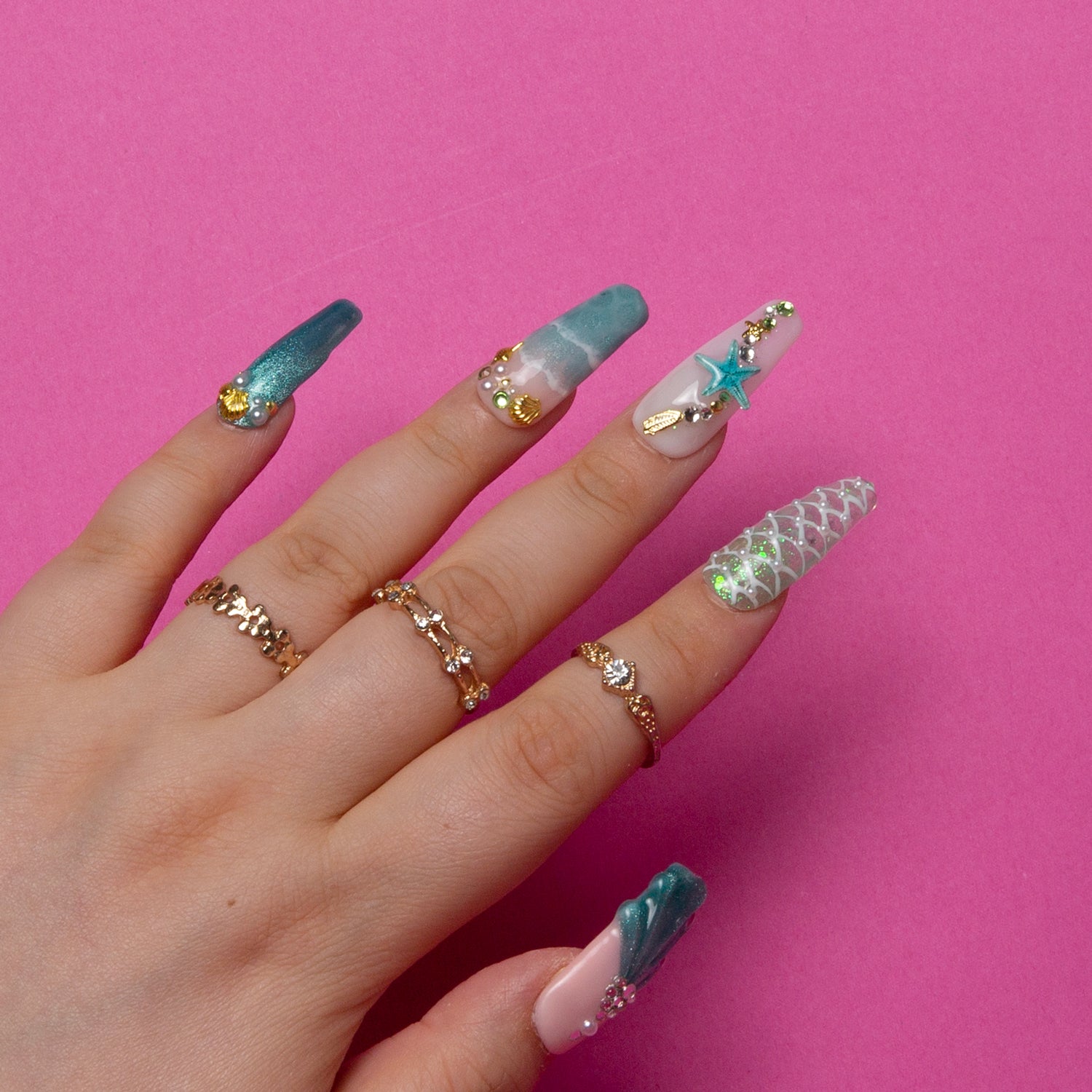 Hand with long coffin-shaped press-on nails in Hawaii sea blue, decorated with starfish and glitter, creating a beach vibe. Gold rings added for elegance. Vibrant pink background.