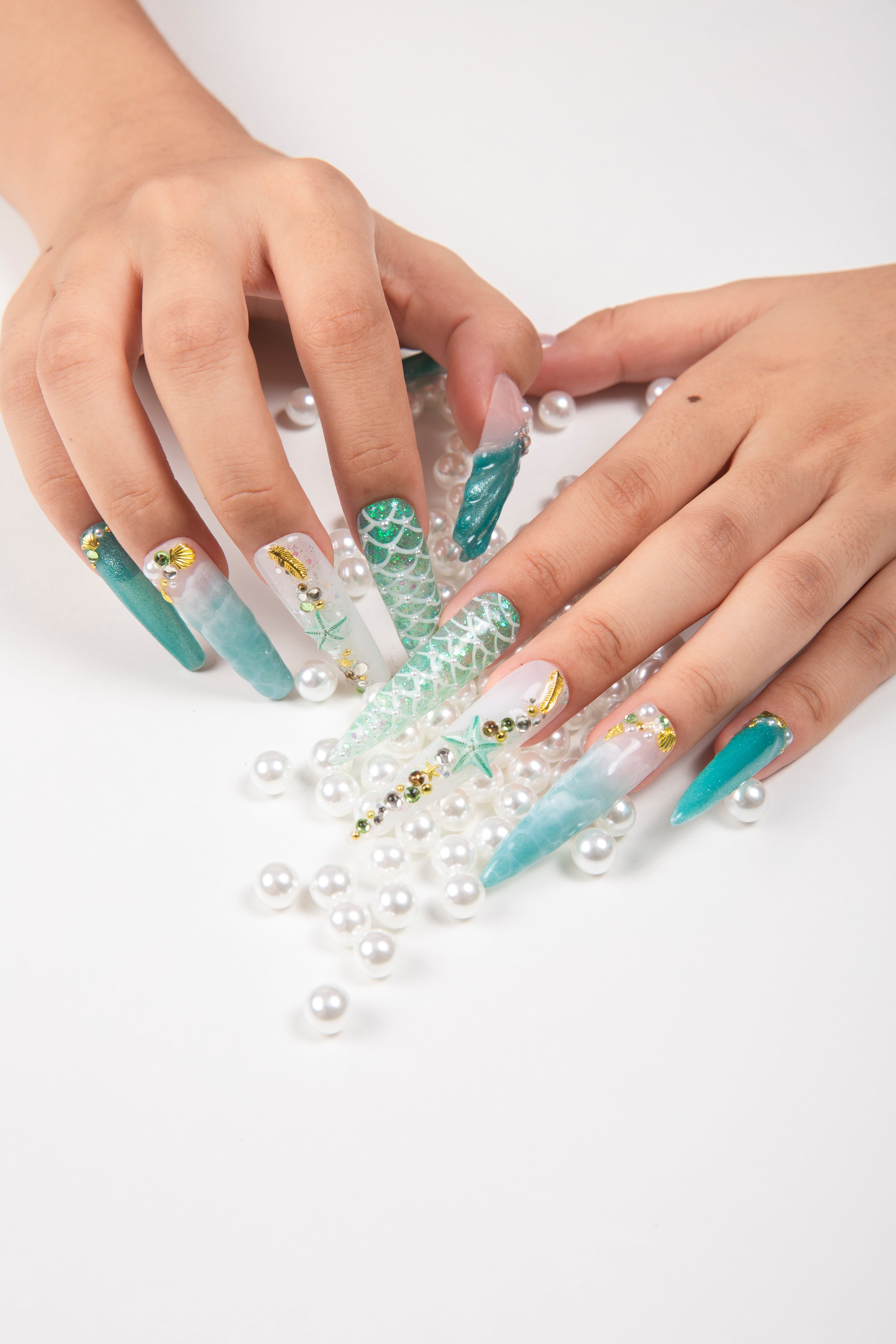 Hands with long press-on nails in Hawaii sea blue theme adorned with starfish, glitter, and intricate designs. Nails are displayed with scattered white pearls, creating a joyful beach vibe.
