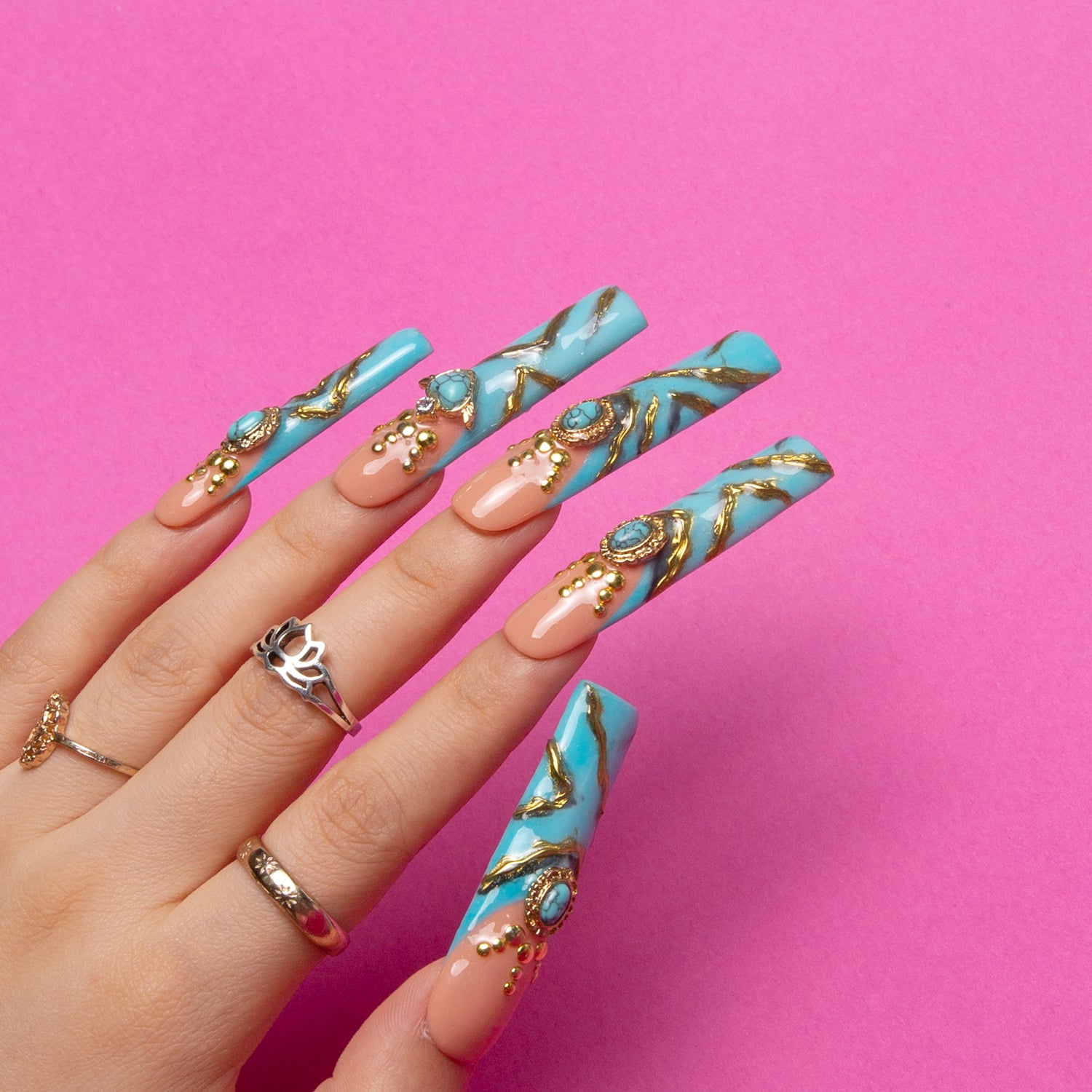 Hand with long, square-shaped turquoise press-on nails featuring golden decor and sparkling gems on a pink background. Rings on fingers enhance the elegant appearance.